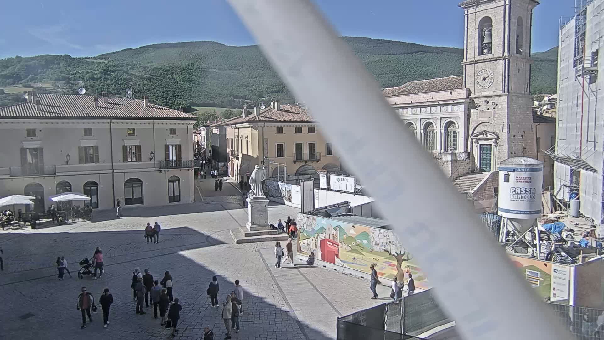 A sunny town square with several people walking around, buildings lining the square, a statue in the center, and construction visible in the background.
