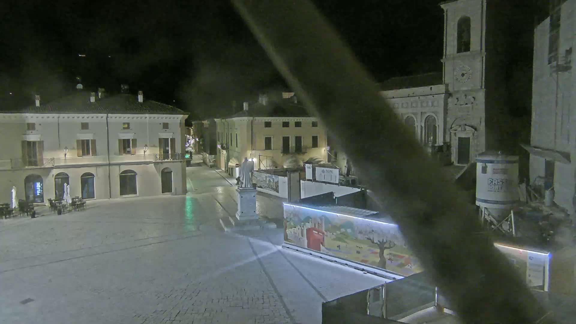 A nighttime view of a town square with several buildings, a statue, and construction materials.