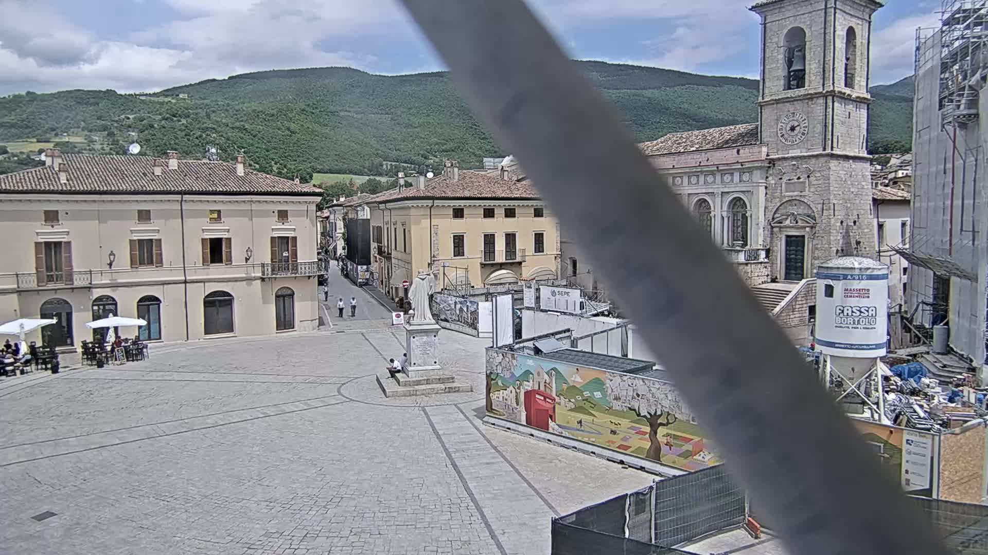 A partly cloudy day reveals a town square with several buildings, a statue, and construction underway, all situated against a backdrop of green hills.