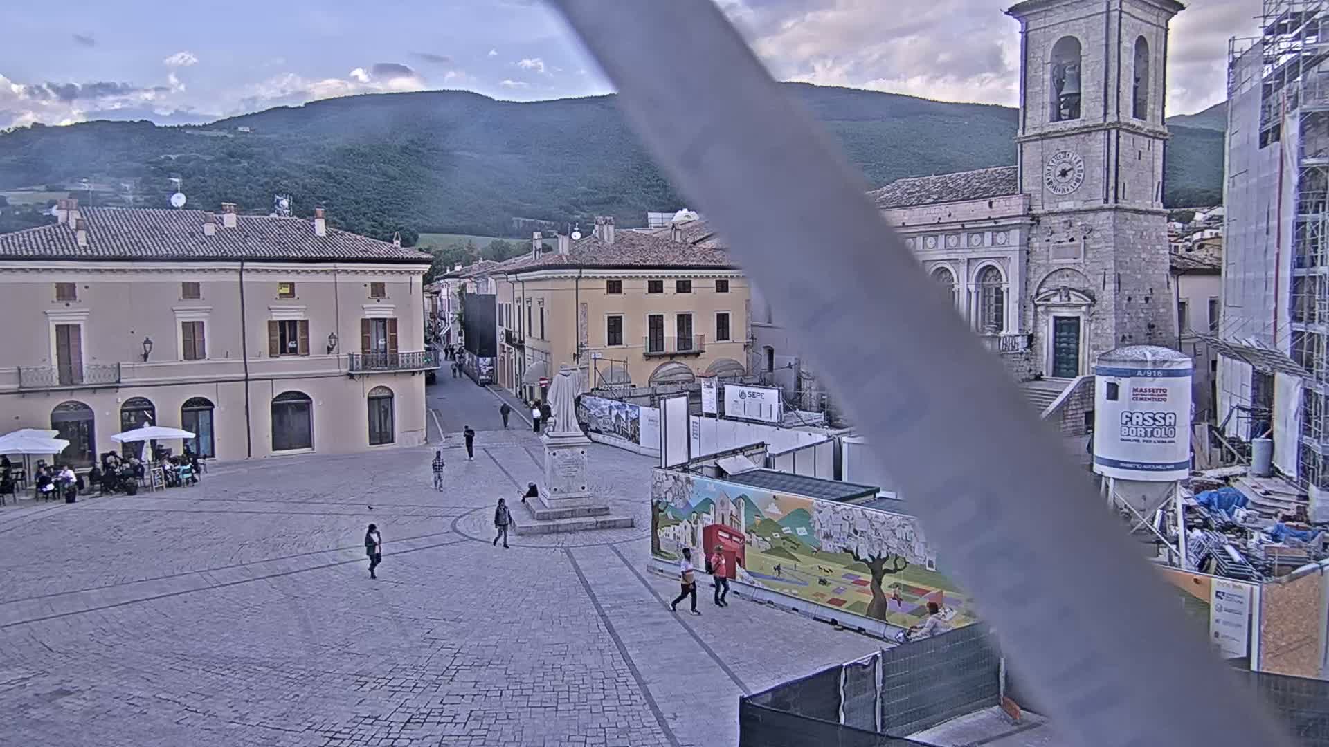A mostly empty town square with several buildings, a statue, and construction in progress, under an overcast sky with mountains in the background.