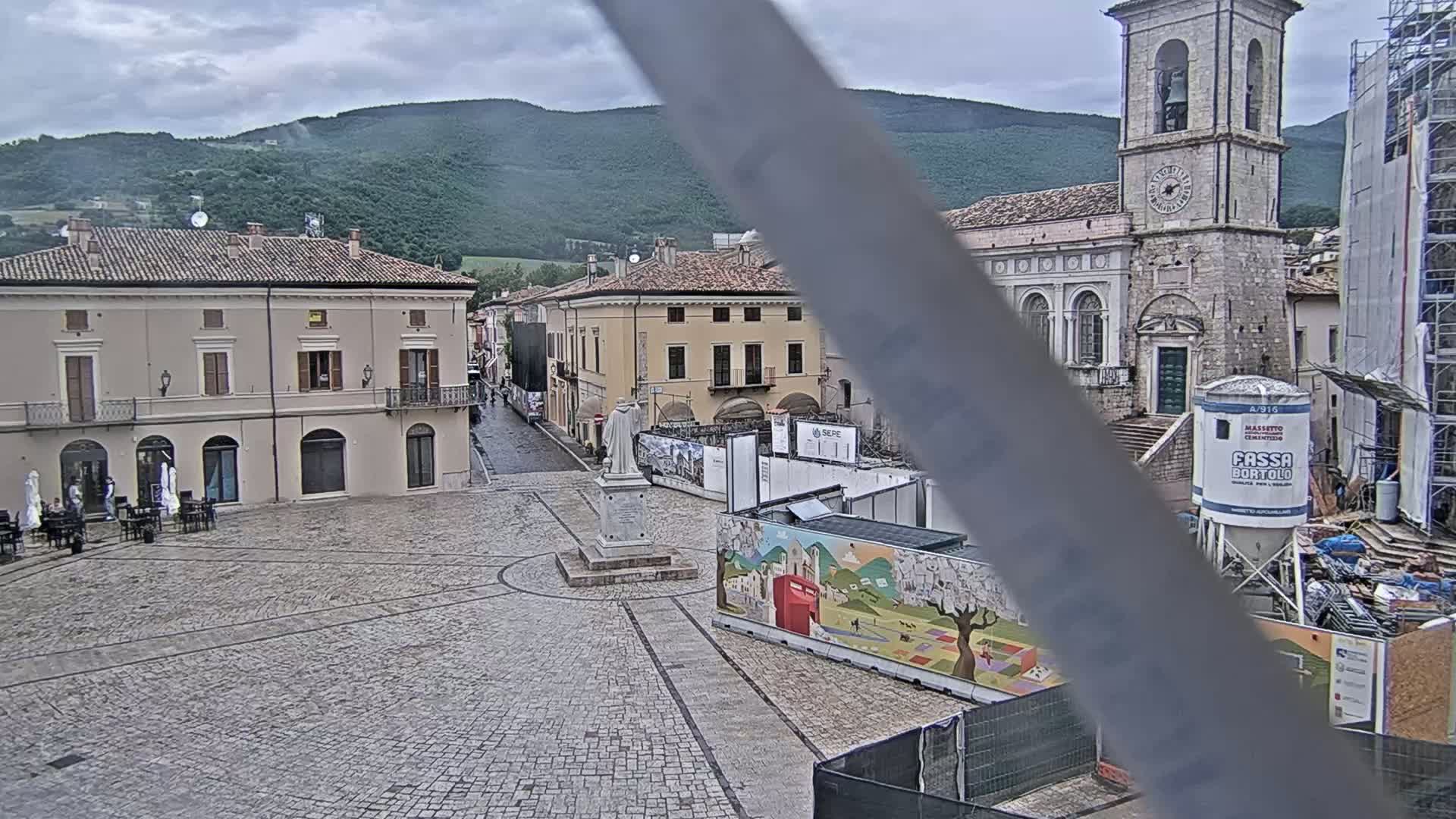 A town square with stone buildings, a statue, and construction underway under an overcast sky.