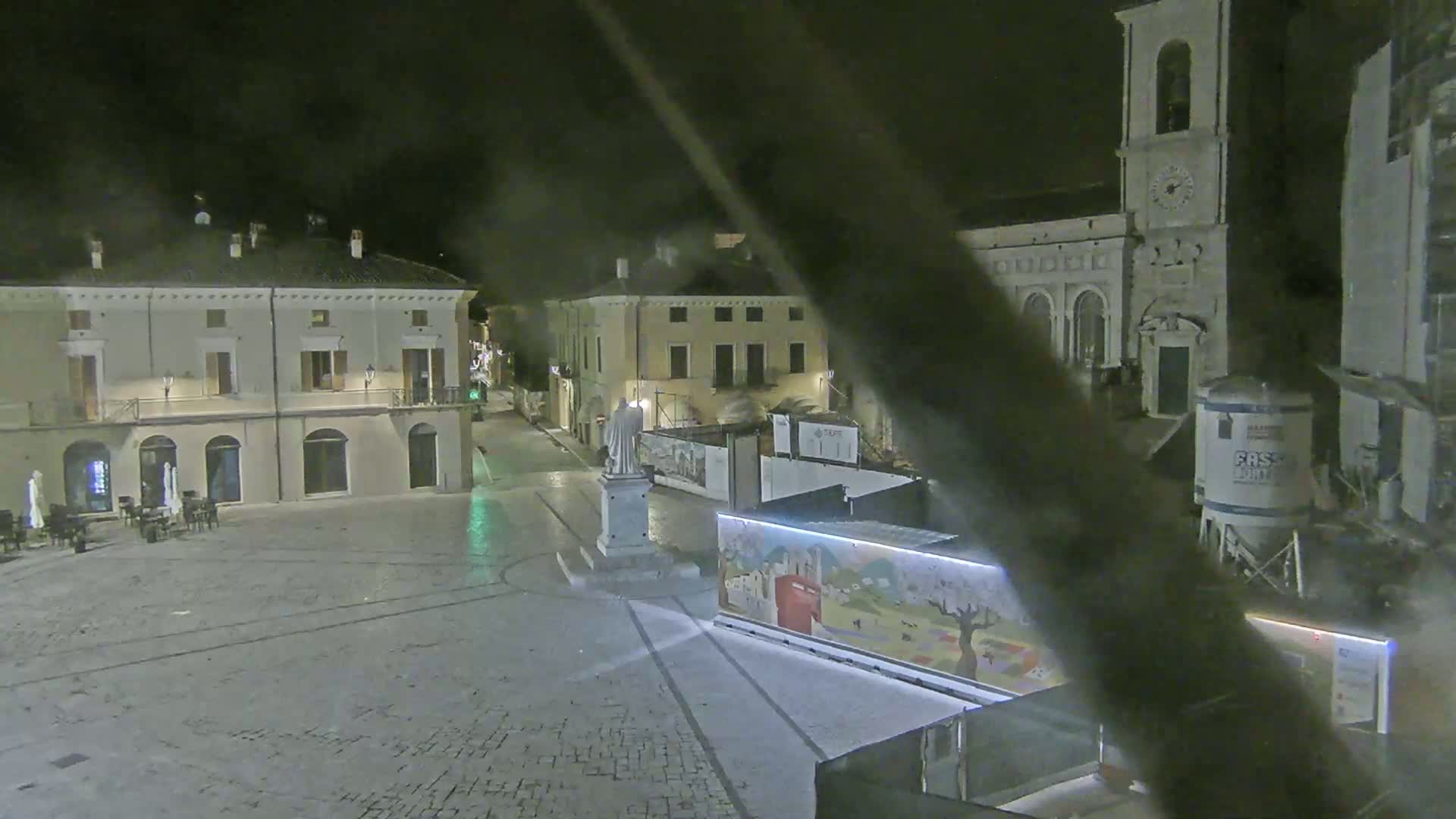 A nighttime view of a town square with buildings, a statue, and construction barriers under a dark sky.
