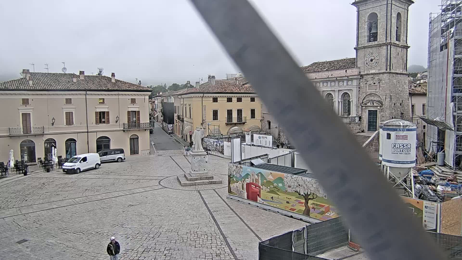 A mostly empty cobblestone town square is visible under an overcast sky, with several buildings, a statue, and some vehicles present, and construction underway in sections of the square.