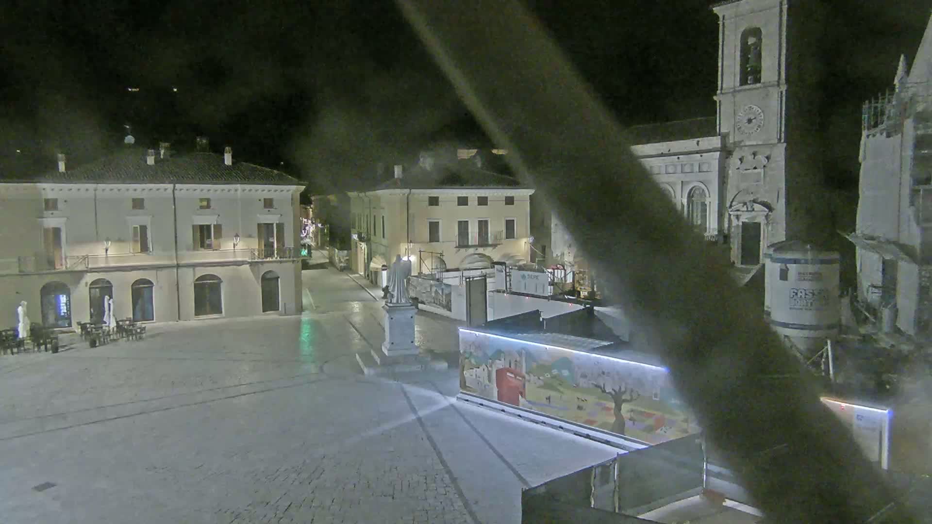 An empty town square at night, with several buildings surrounding it, including a church with a tall bell tower, and construction or renovation taking place near a statue.