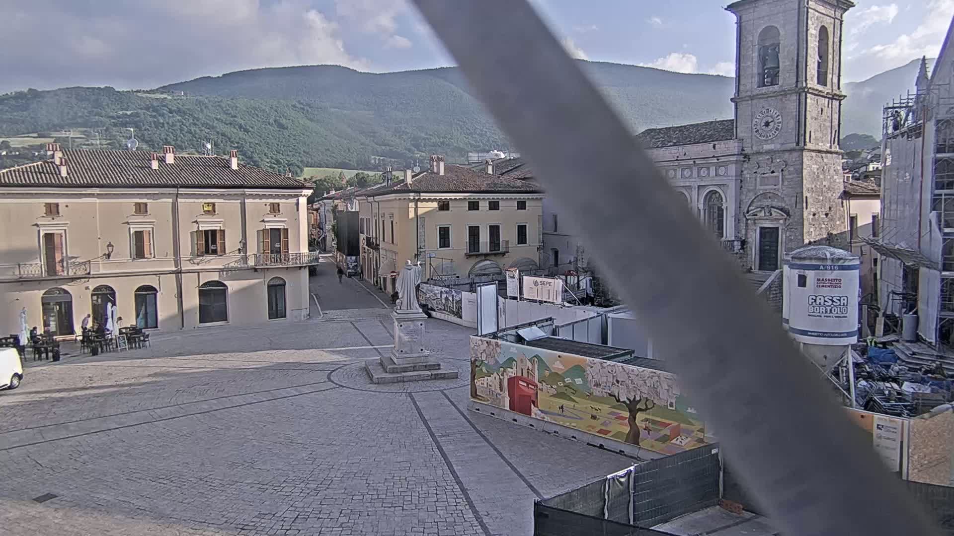 A town square with several buildings, a statue, and construction underway, all under a partly cloudy sky, with mountains visible in the background.