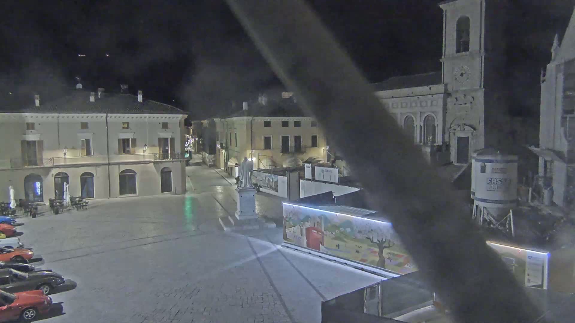 A nighttime view of a town square with several buildings, a statue, and cars parked along one side, under dark, clear skies.