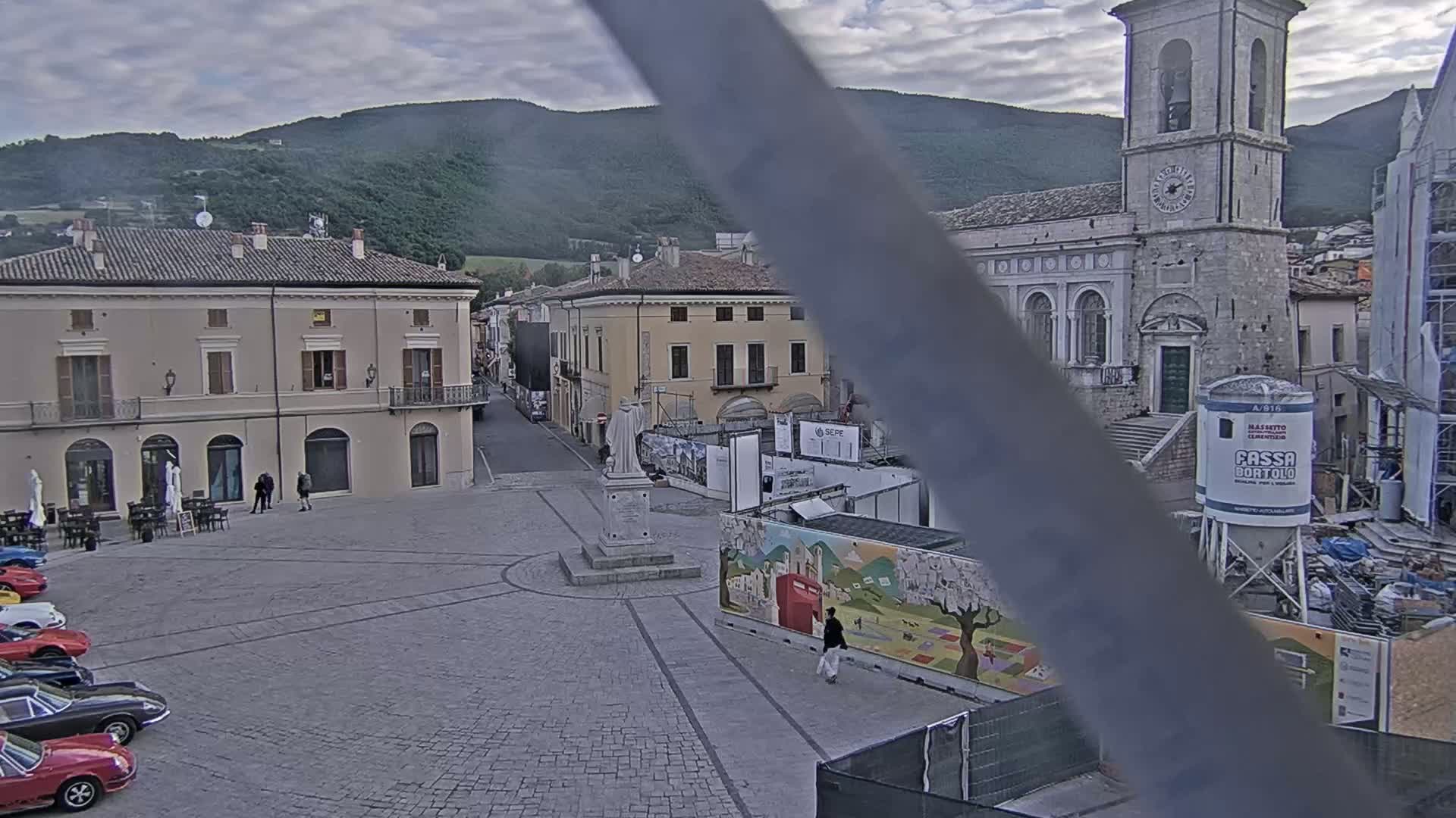 A town square with several parked cars, a few pedestrians, and buildings under a cloudy sky, with a church and mountains visible in the background.