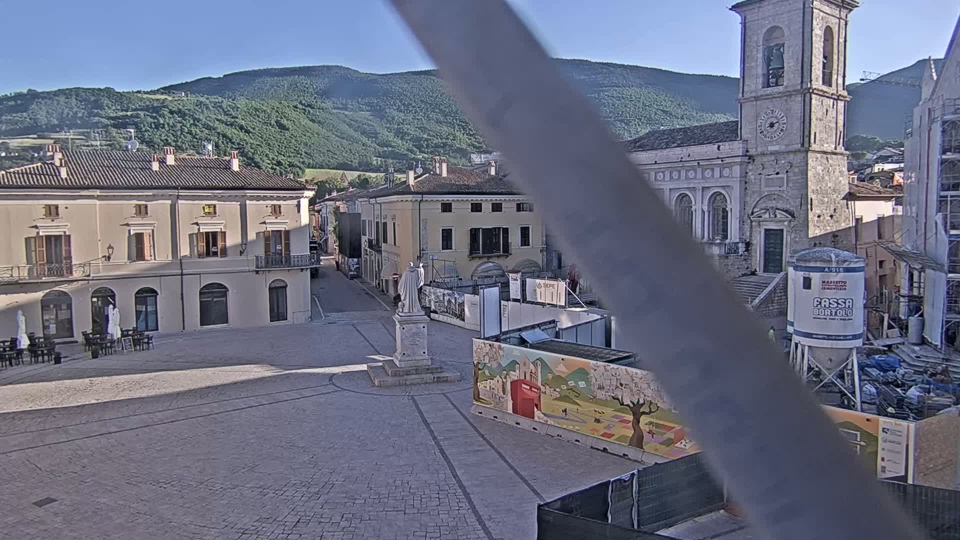 A town square in a mountainous region on a clear, sunny day, featuring a statue, several buildings, and construction work in progress.