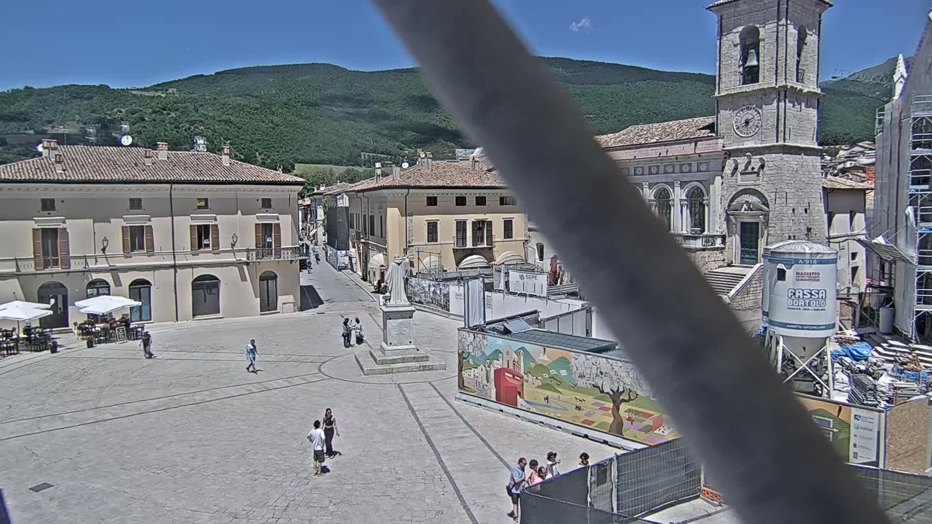 A town square with several buildings, a statue, and some people is seen under sunny skies, with mountains in the background and construction underway in part of the square.