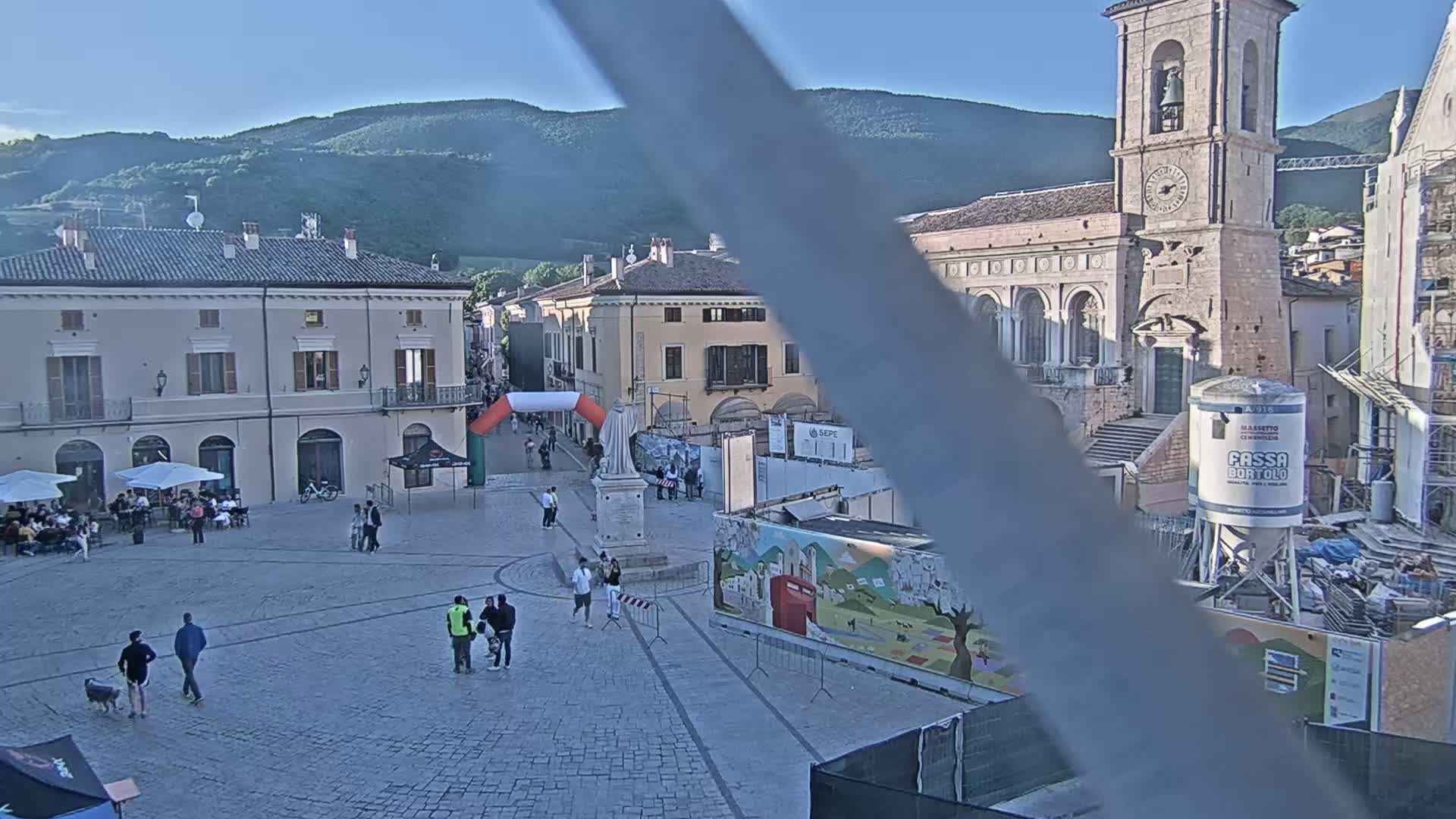 A town square, under sunny skies, features several people walking around, buildings lining the perimeter, and construction underway in a section of the square.