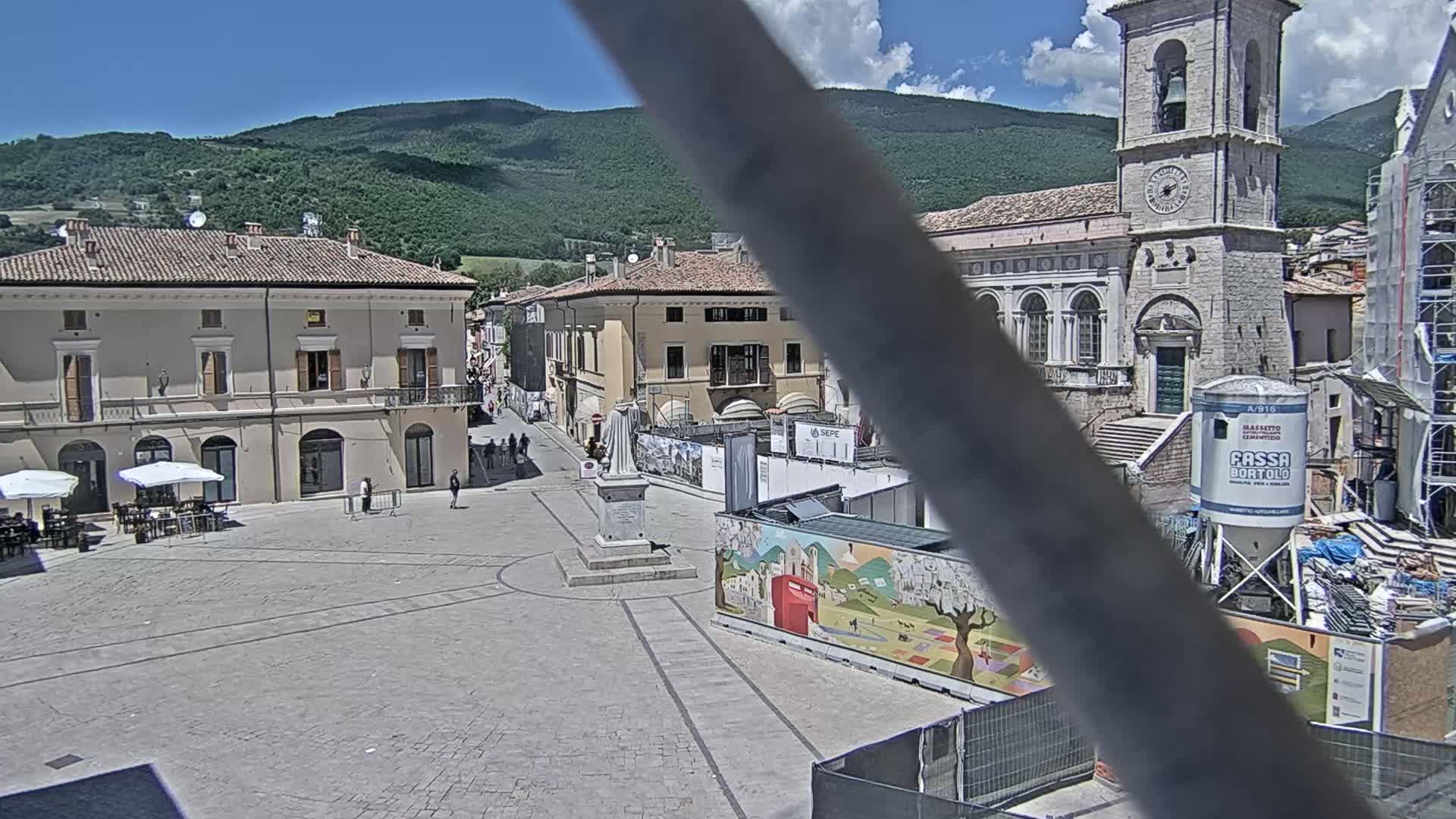 A town square in a European-style town, under a sunny sky, is partially obstructed by construction and features buildings with classical architecture and a tall bell tower.