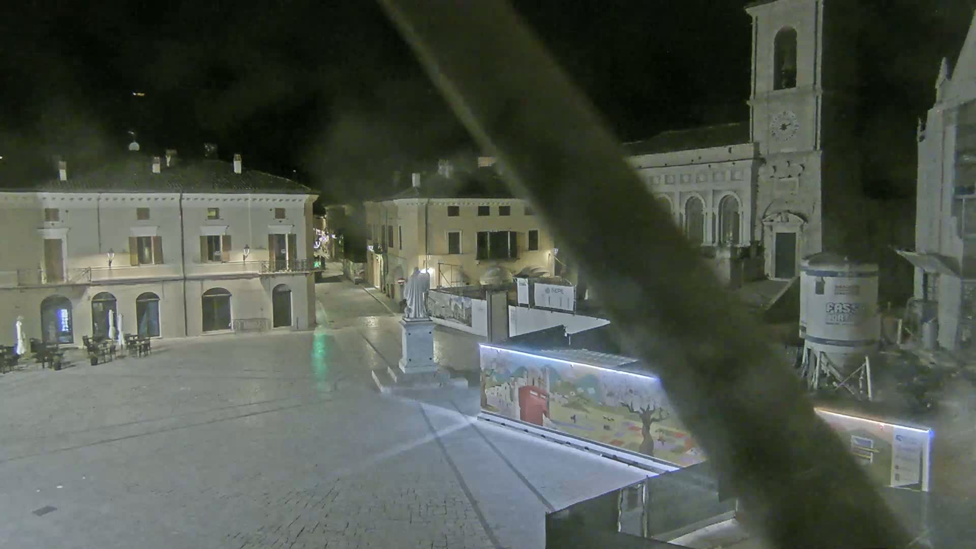 A nighttime view of a town square with several buildings, a statue, and construction materials, under clear skies.