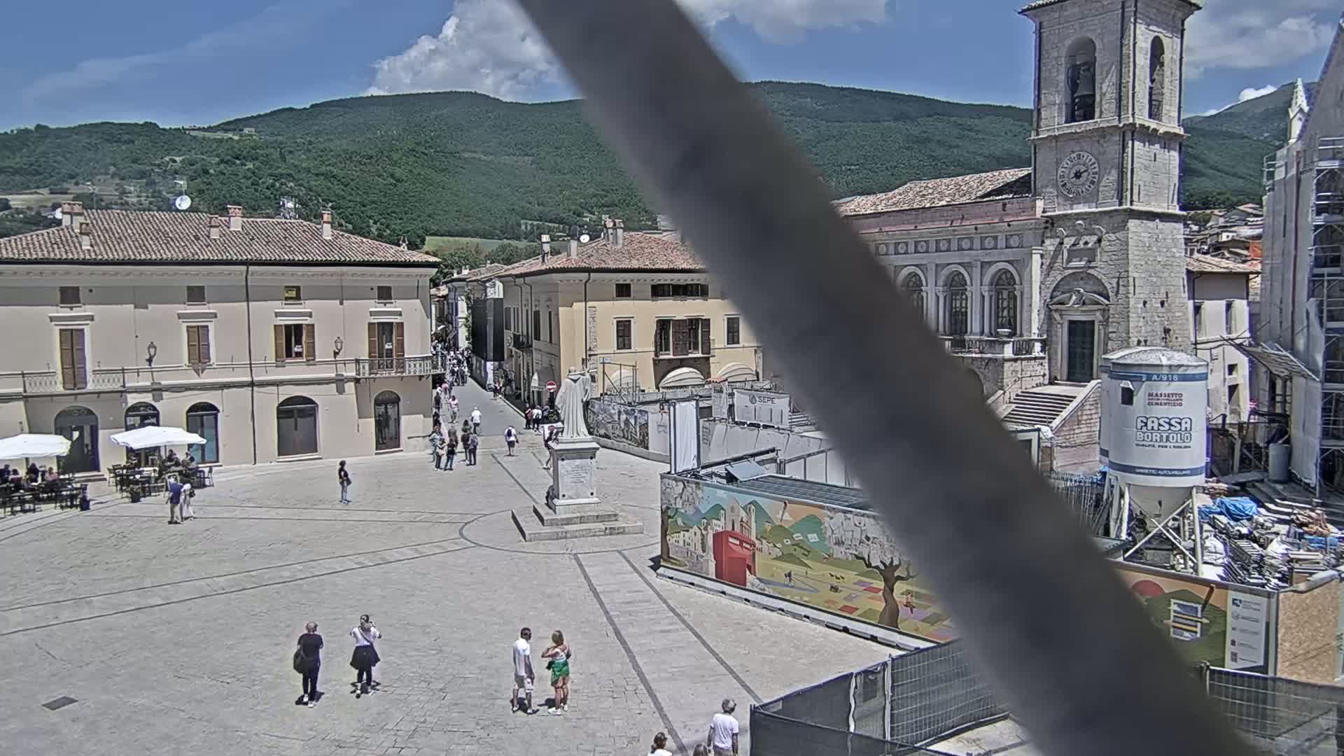 A town square with several buildings, a statue, and a few people, under a sunny sky with mountains in the background.
