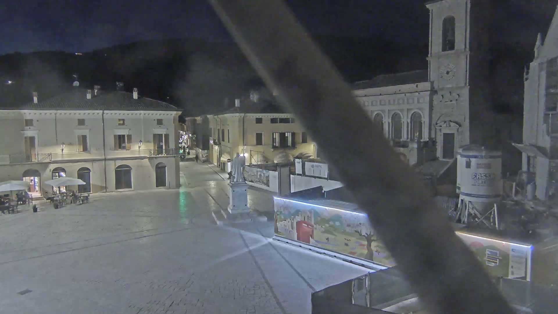 A nighttime view of a town square with several buildings, a statue, and some outdoor seating under the cover of darkness.