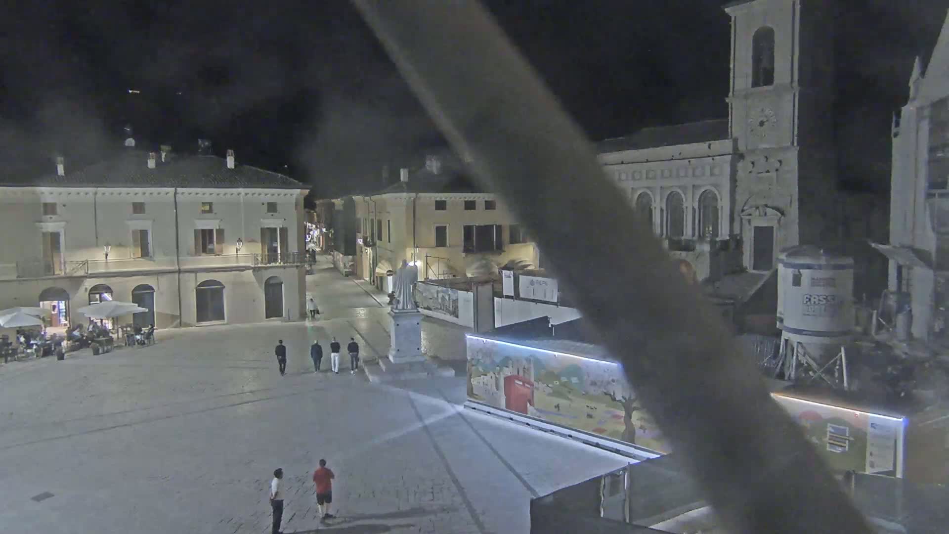 A nighttime view of a town square, with several buildings surrounding it, a statue in the center, and a few people walking around.