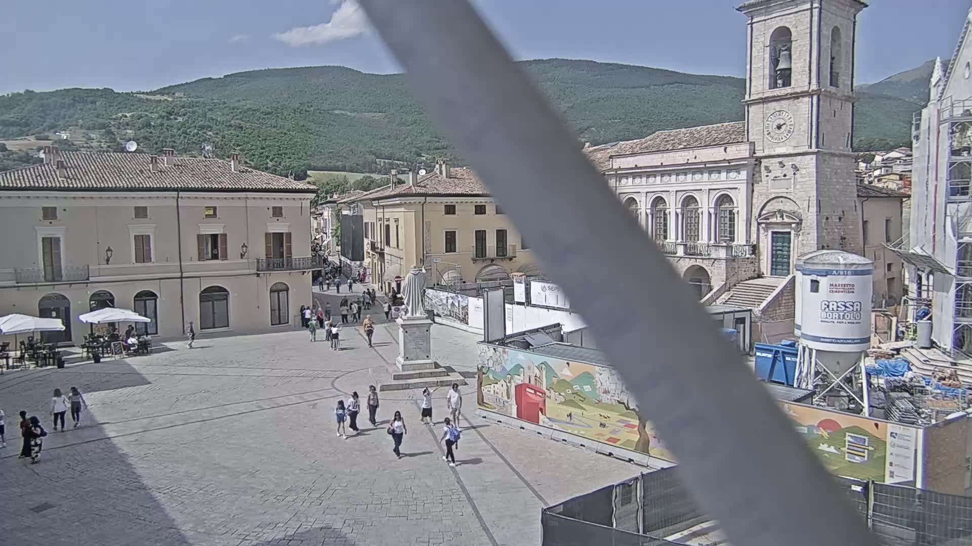 A town square with several buildings, some under construction, is populated with a few people on a sunny day.