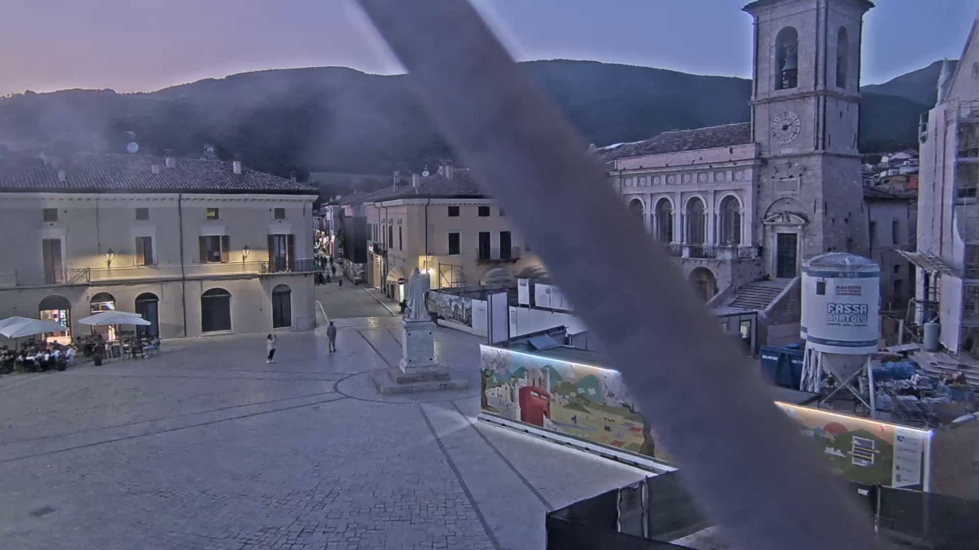 A town square at dusk, under a mostly clear sky, features buildings, a statue, and construction equipment.