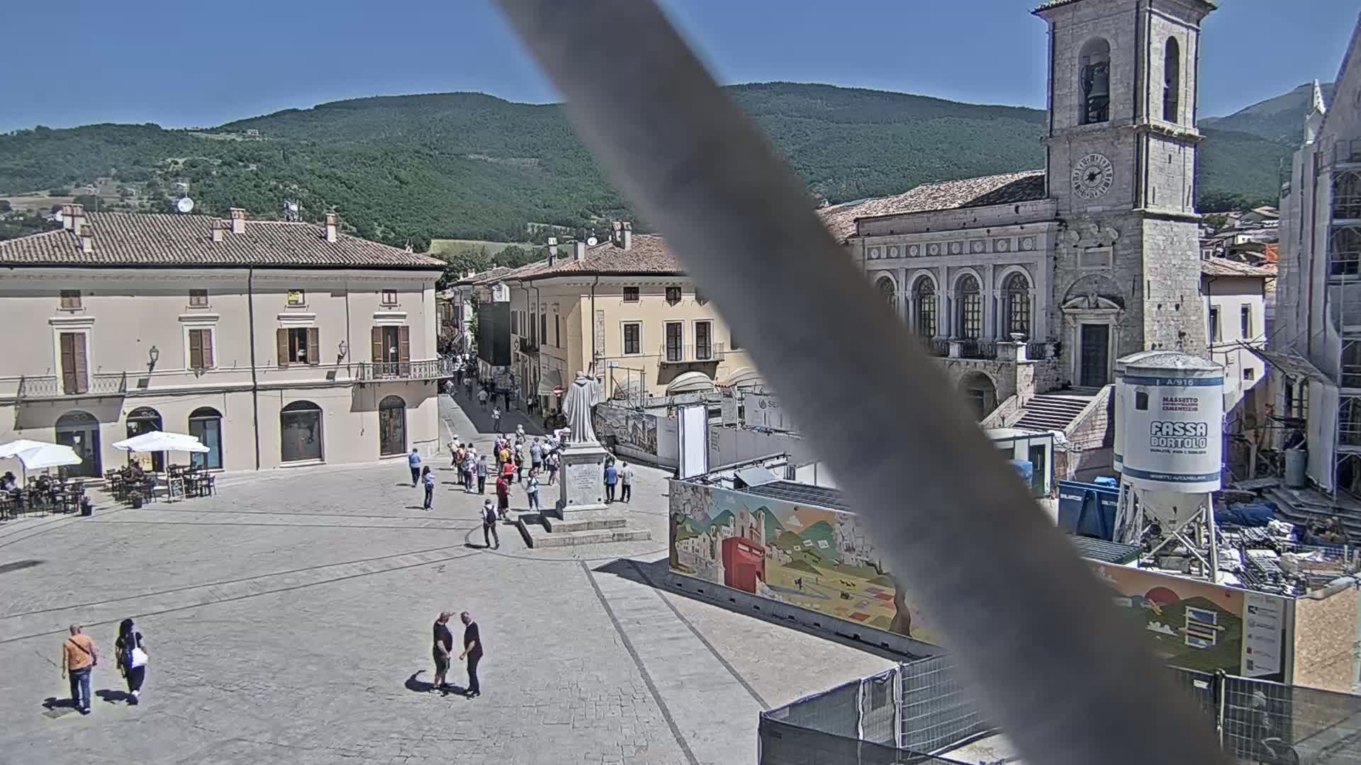 A sunny town square, surrounded by buildings, with a few people walking around and some construction taking place.