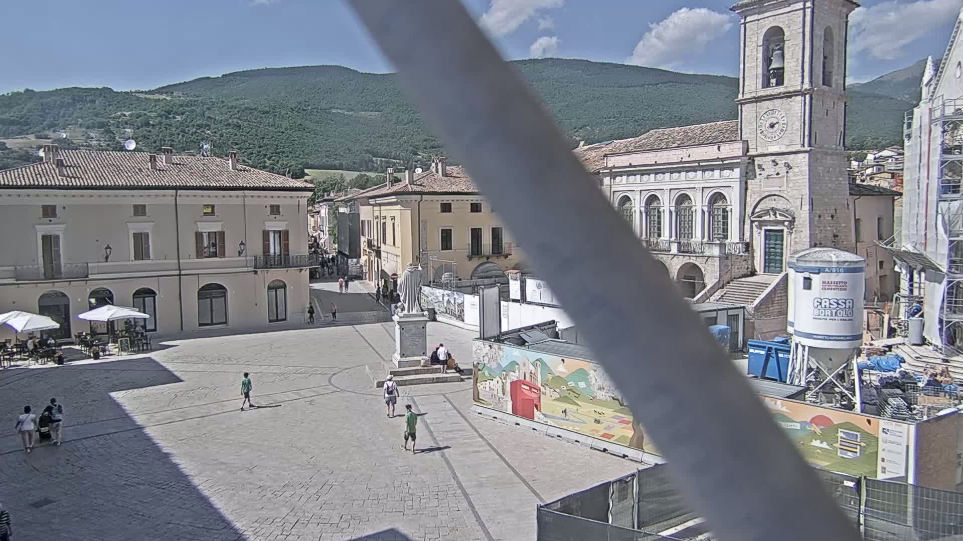 A town square with several buildings, a statue, and a few people, under a sunny sky with mountains in the background.