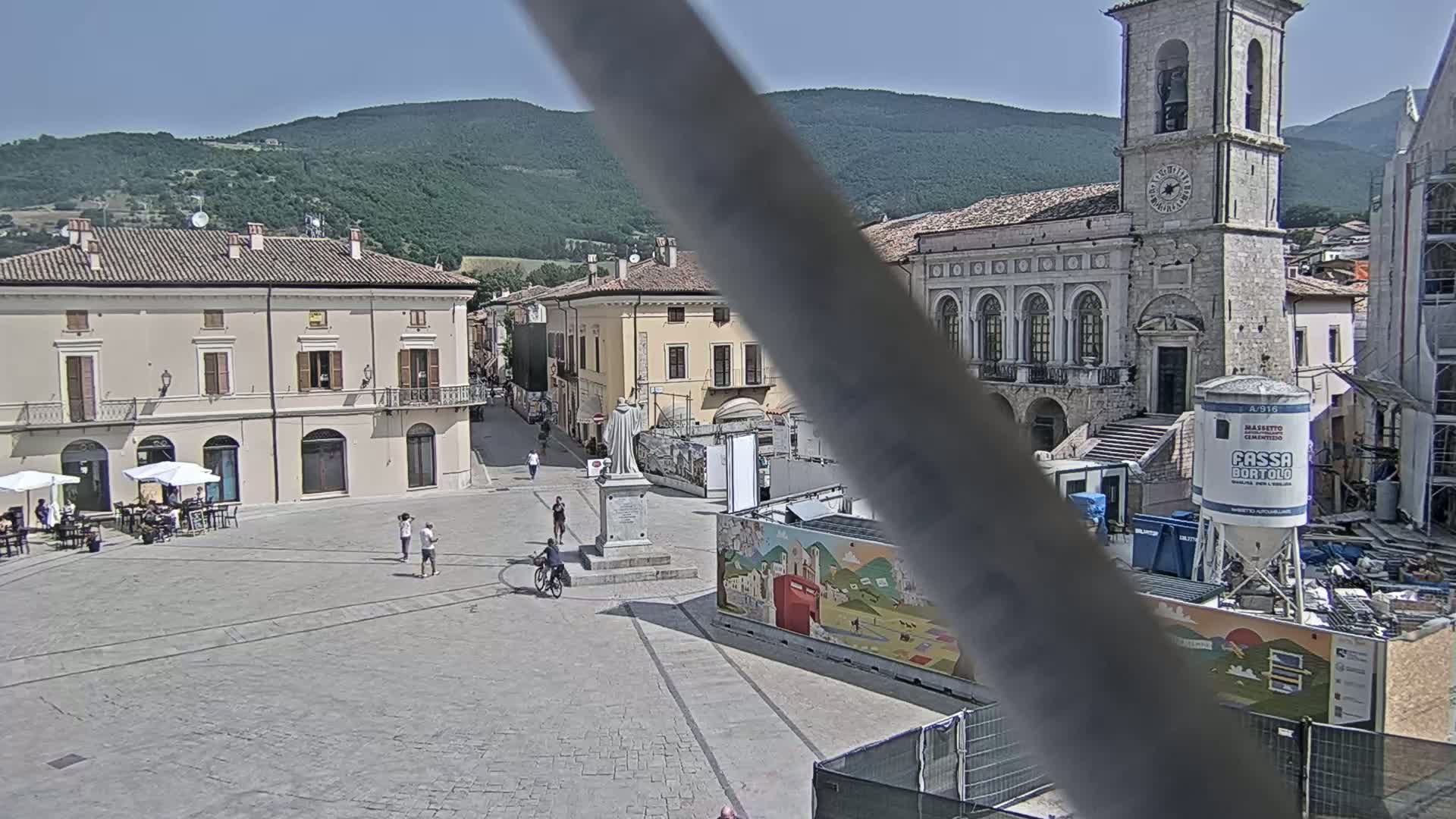A town square with several buildings, a monument, and a few people, under a sunny sky and against a backdrop of green hills.