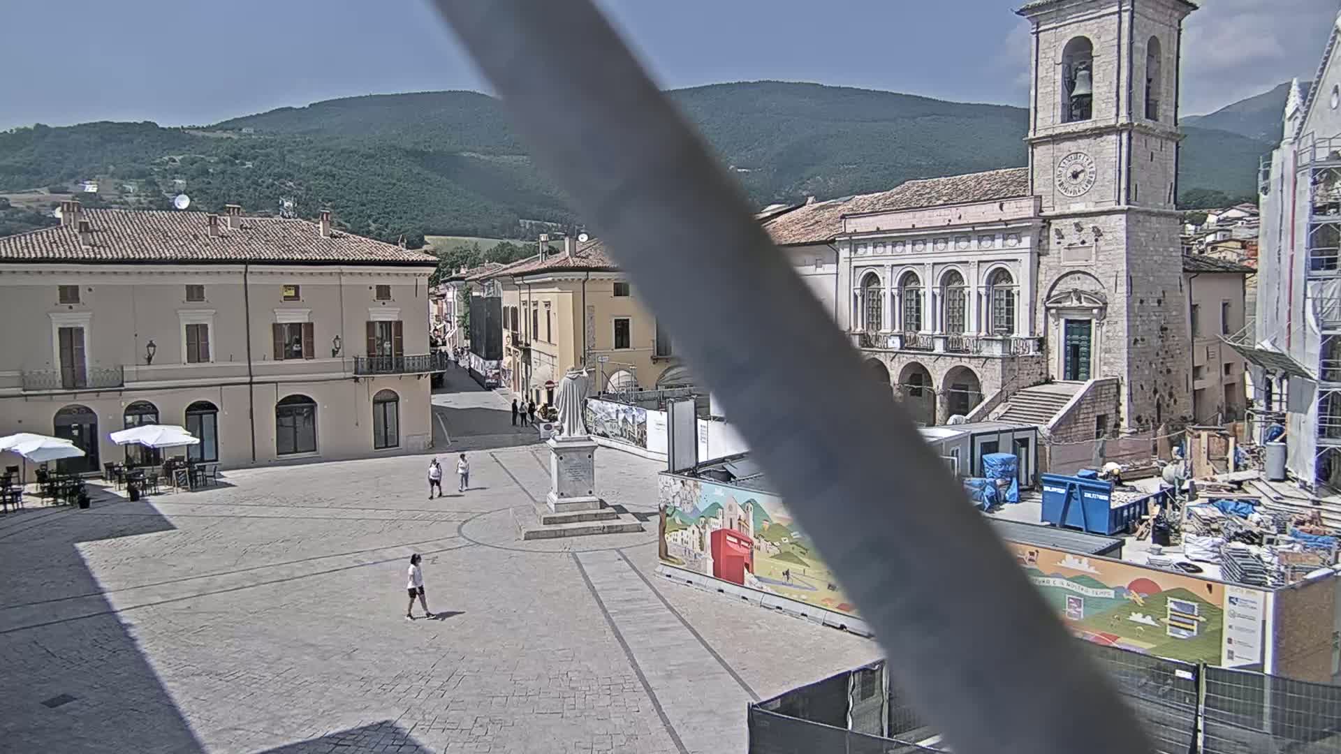 A town square with several buildings, a statue, and construction underway, all under a clear sunny sky and backed by a mountain range.