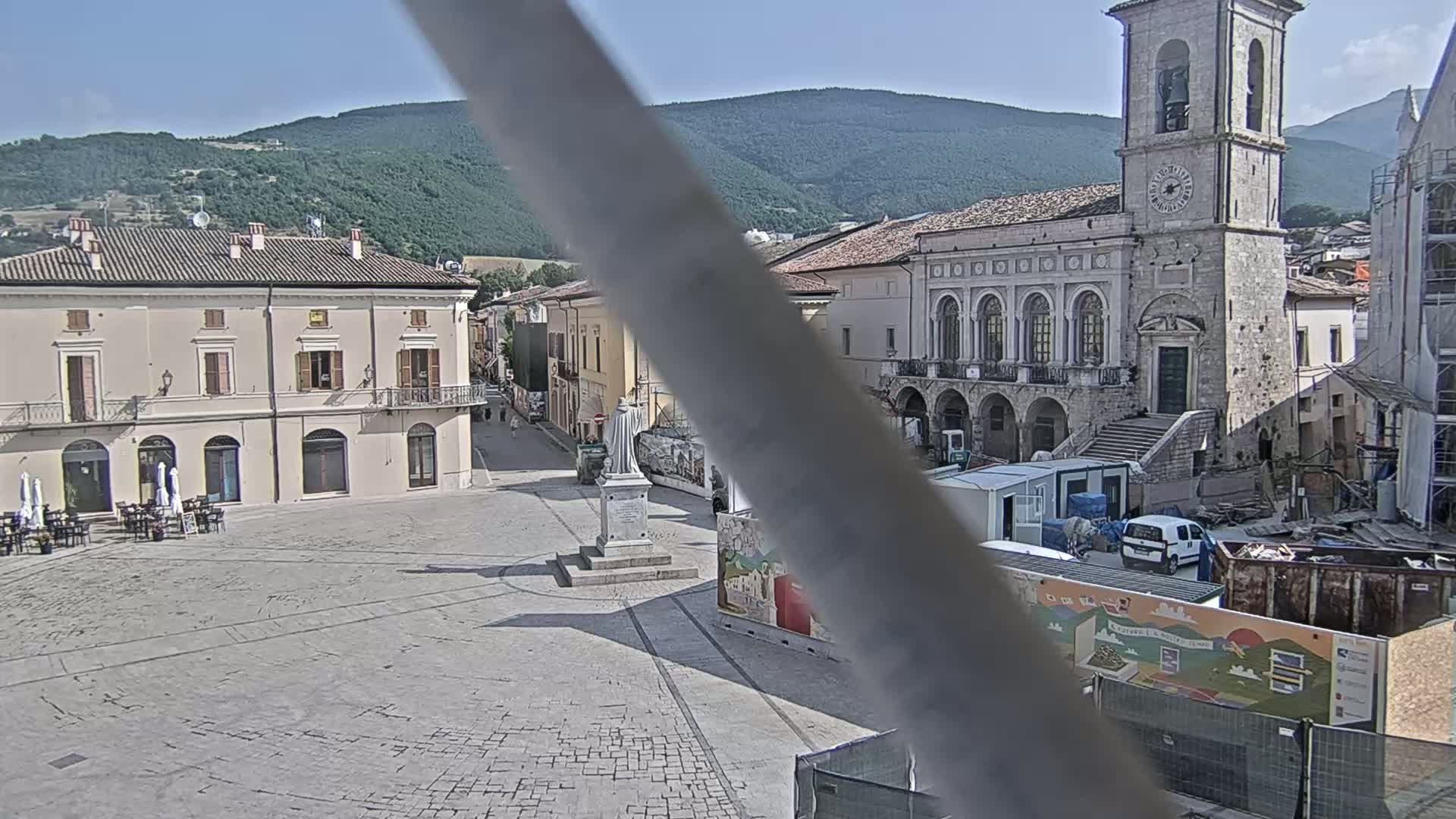 A town square with several buildings, a statue, and construction equipment is visible on a sunny day, with mountains in the background.