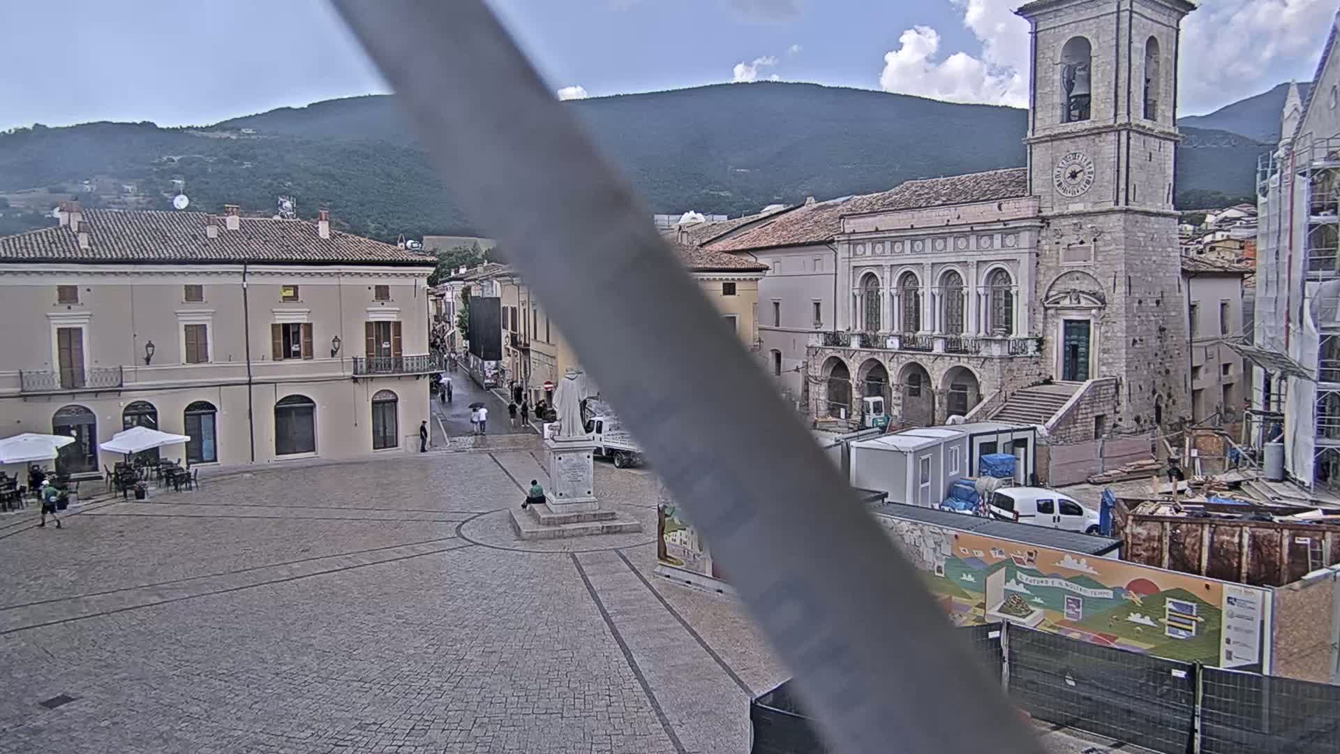A town square with stone buildings, including a tall clock tower, under a partly cloudy sky, with some construction or renovation taking place in the square.