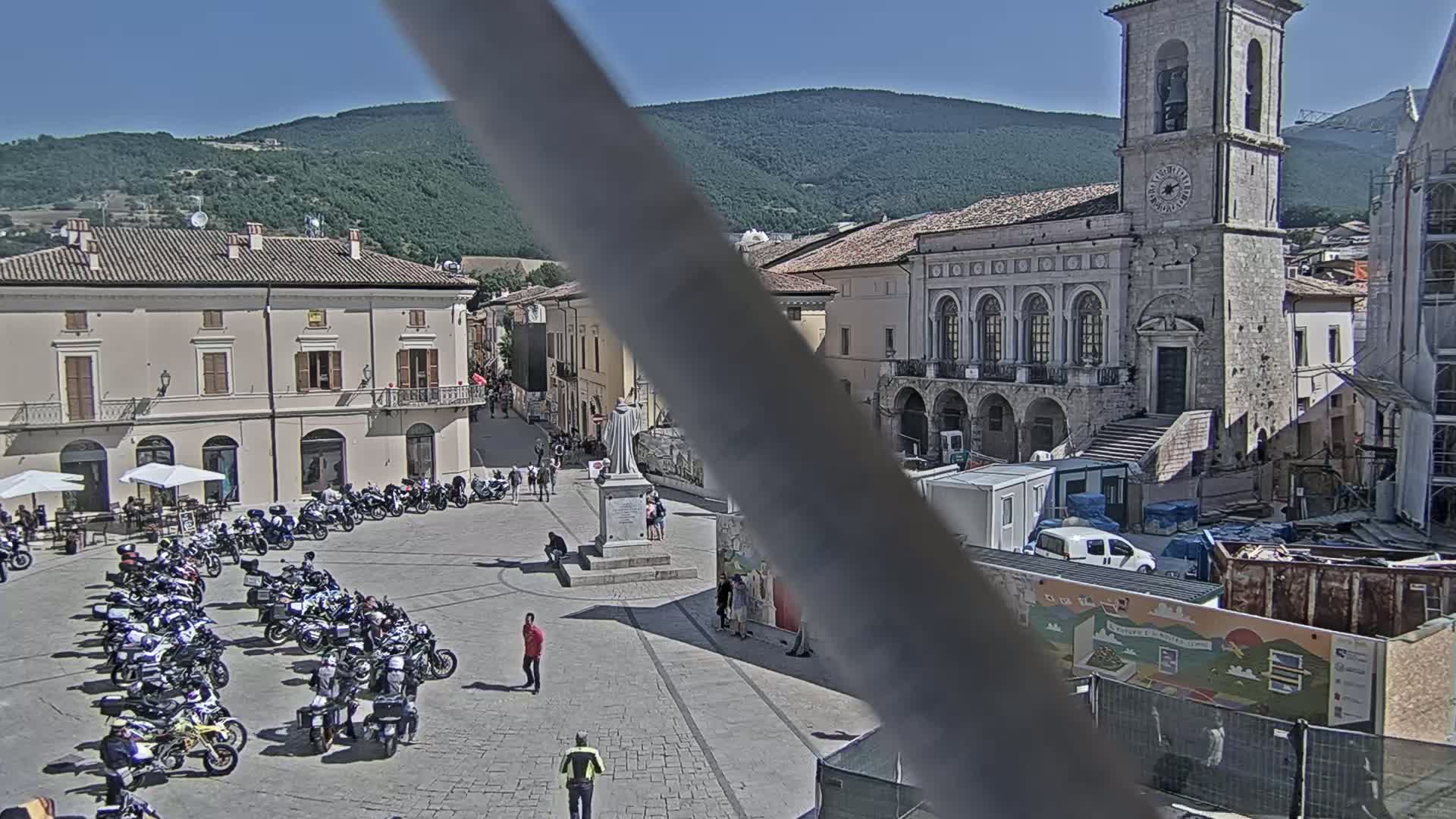 A town square filled with parked motorcycles, a monument, and several buildings under a sunny sky.
