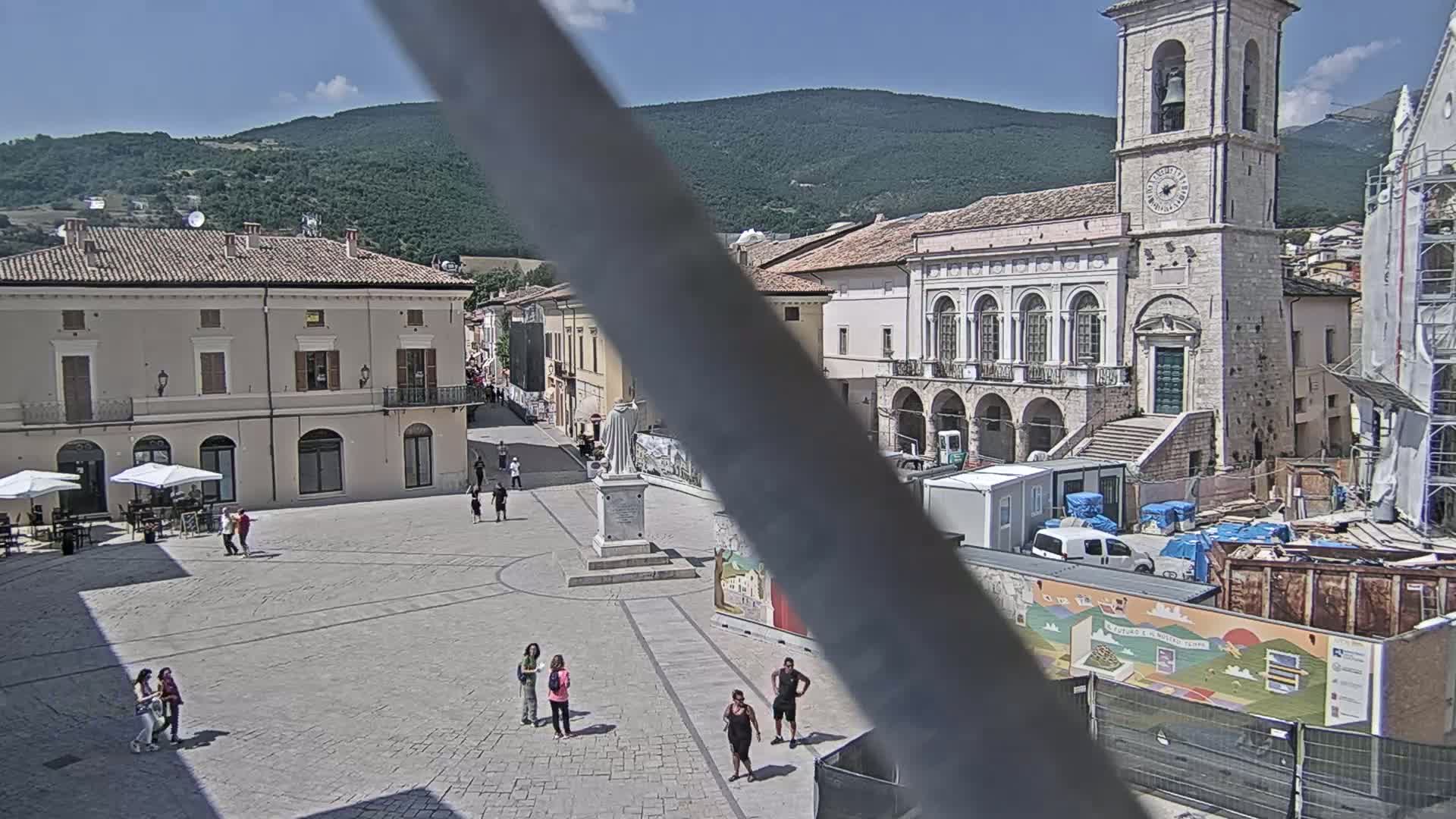A town square with several buildings, a statue, and a few people, under sunny skies and in front of a mountain range.