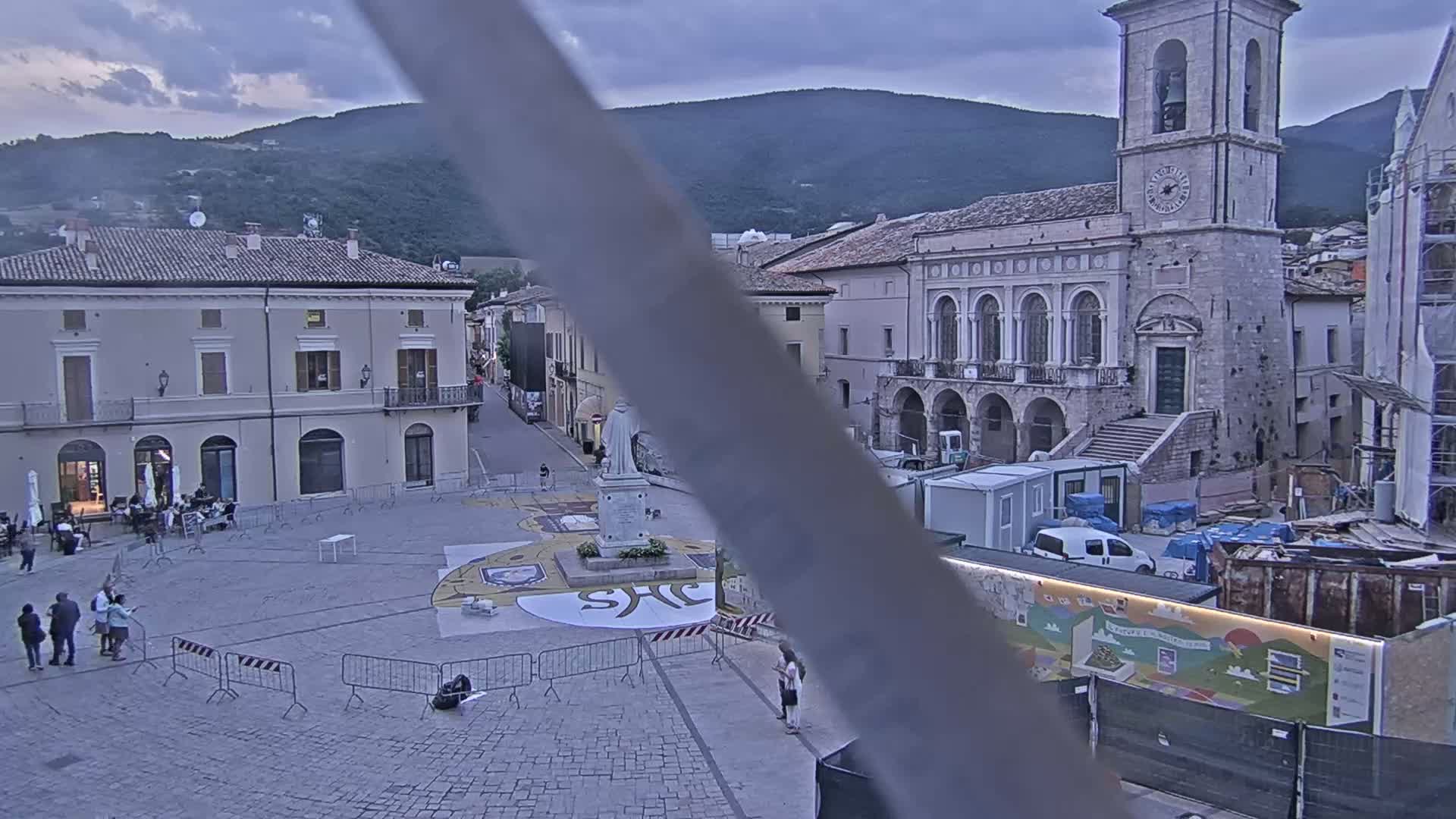 A town square with several buildings, a statue, and some people, under an overcast sky, is partially obscured by a large vertical object.