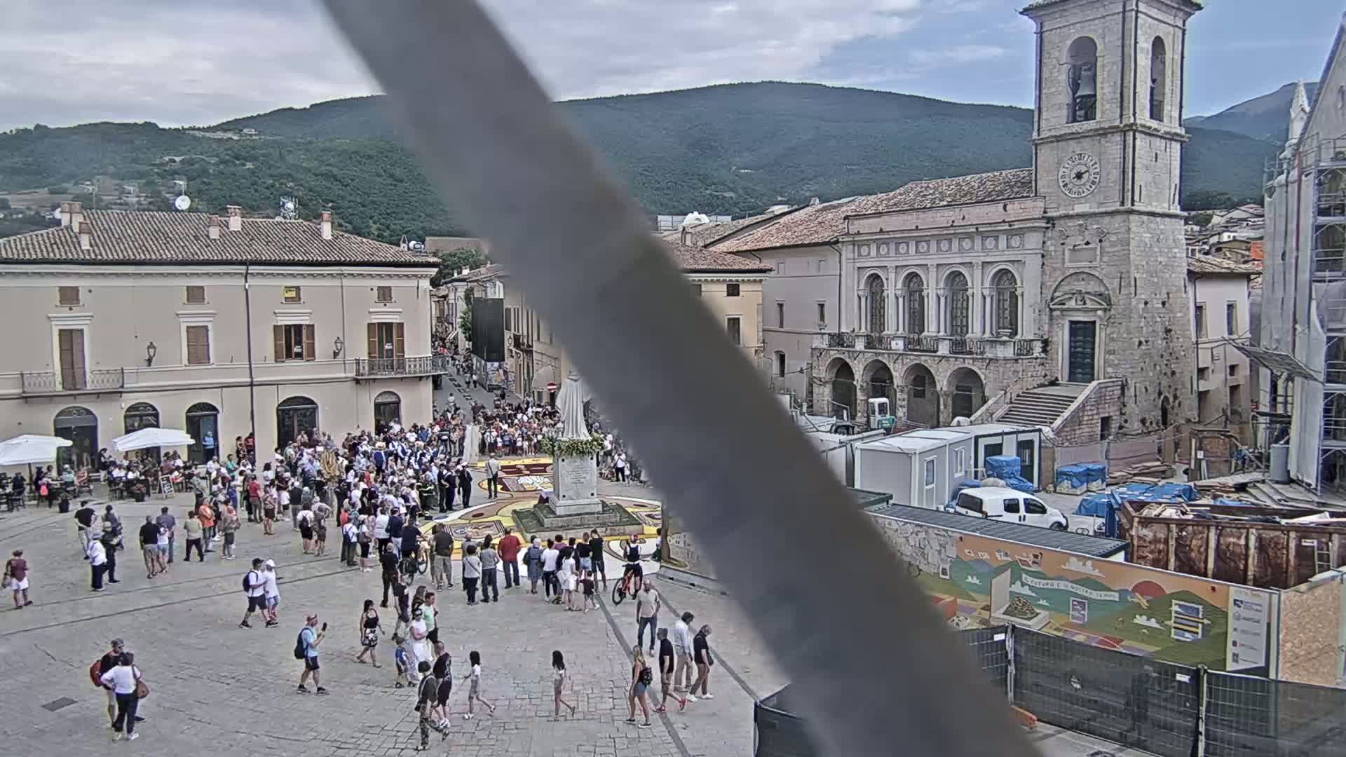 A town square filled with people, centered around a large, colorful design on the ground, is surrounded by buildings, including a tall stone church, under a mostly cloudy sky.