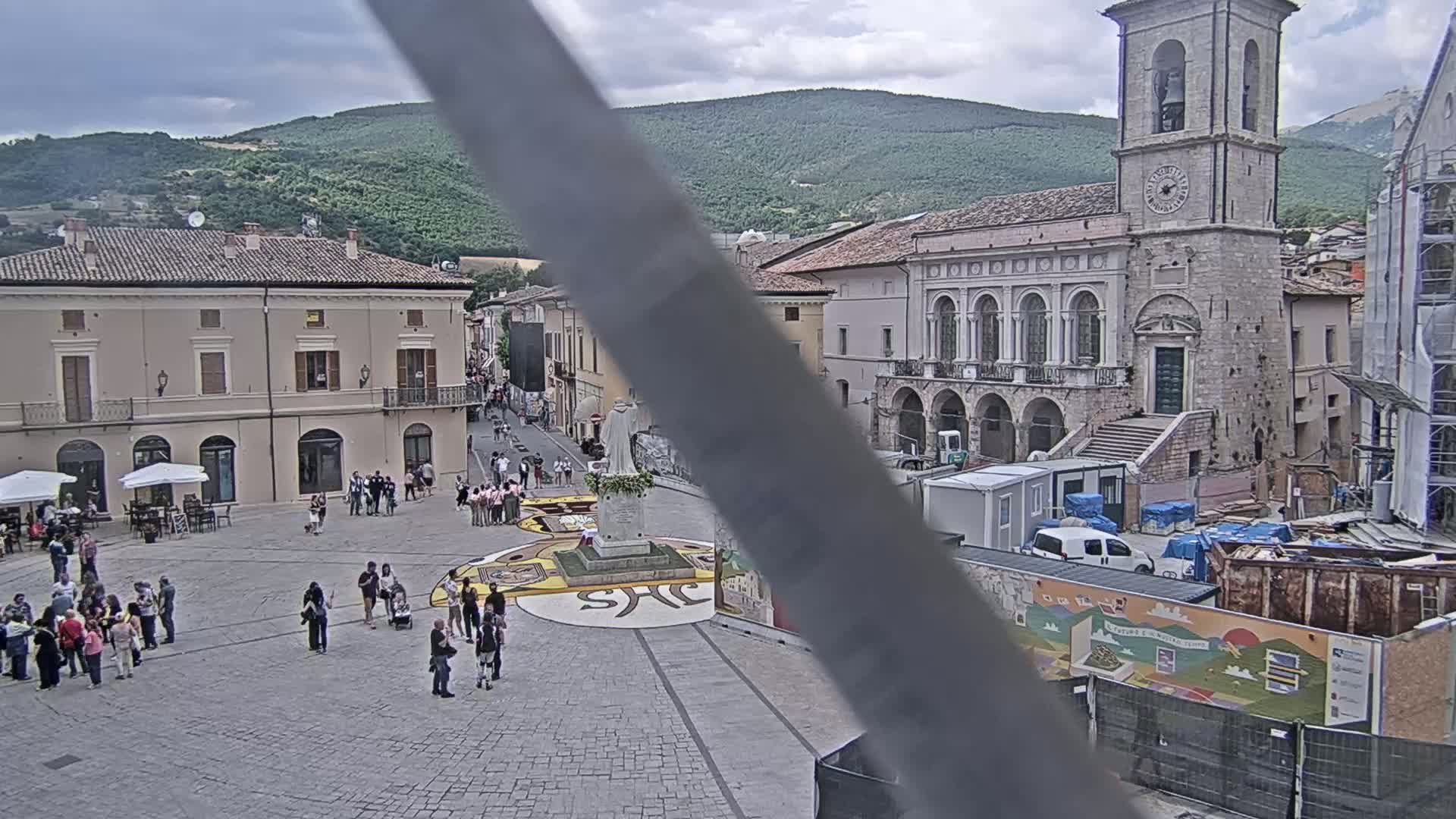 A town square with people gathered around a large, decorated circular area in front of historical buildings under an overcast sky, with mountains visible in the background.