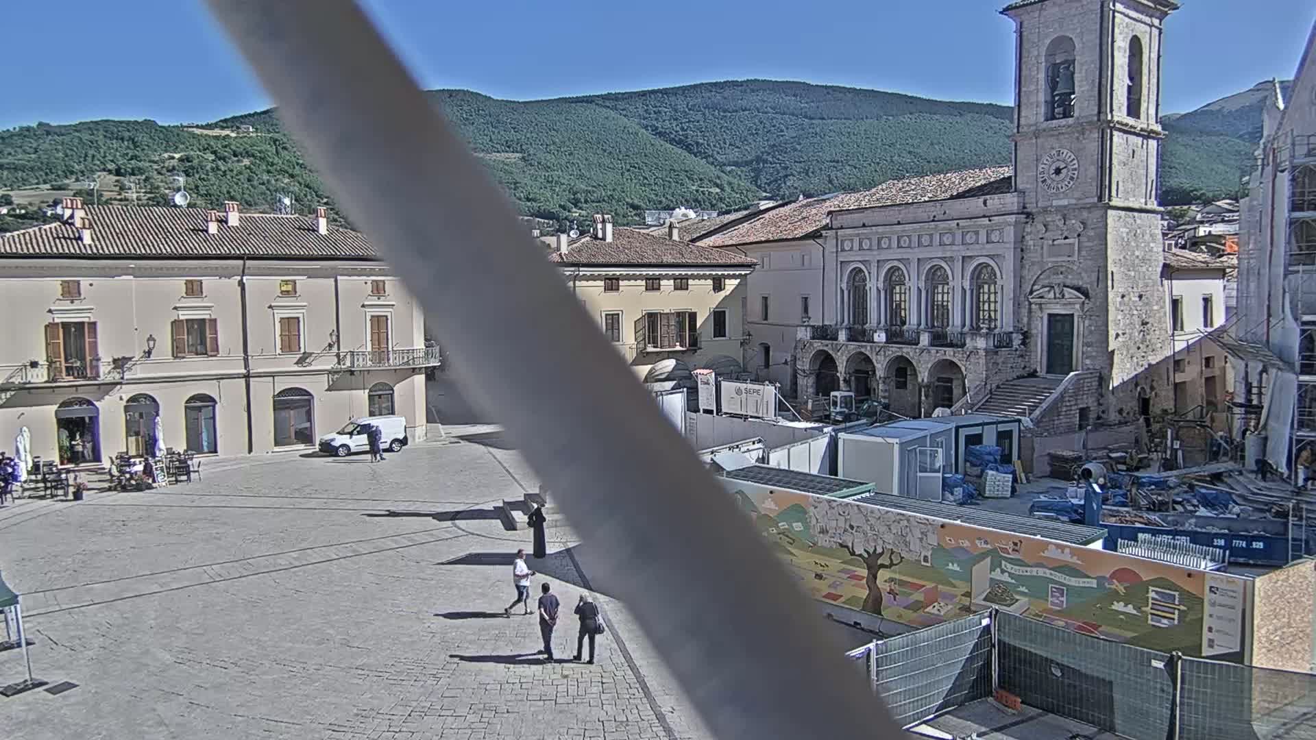 A town square with several stone buildings, some under construction, is situated against a backdrop of green hills under a clear, sunny sky.
