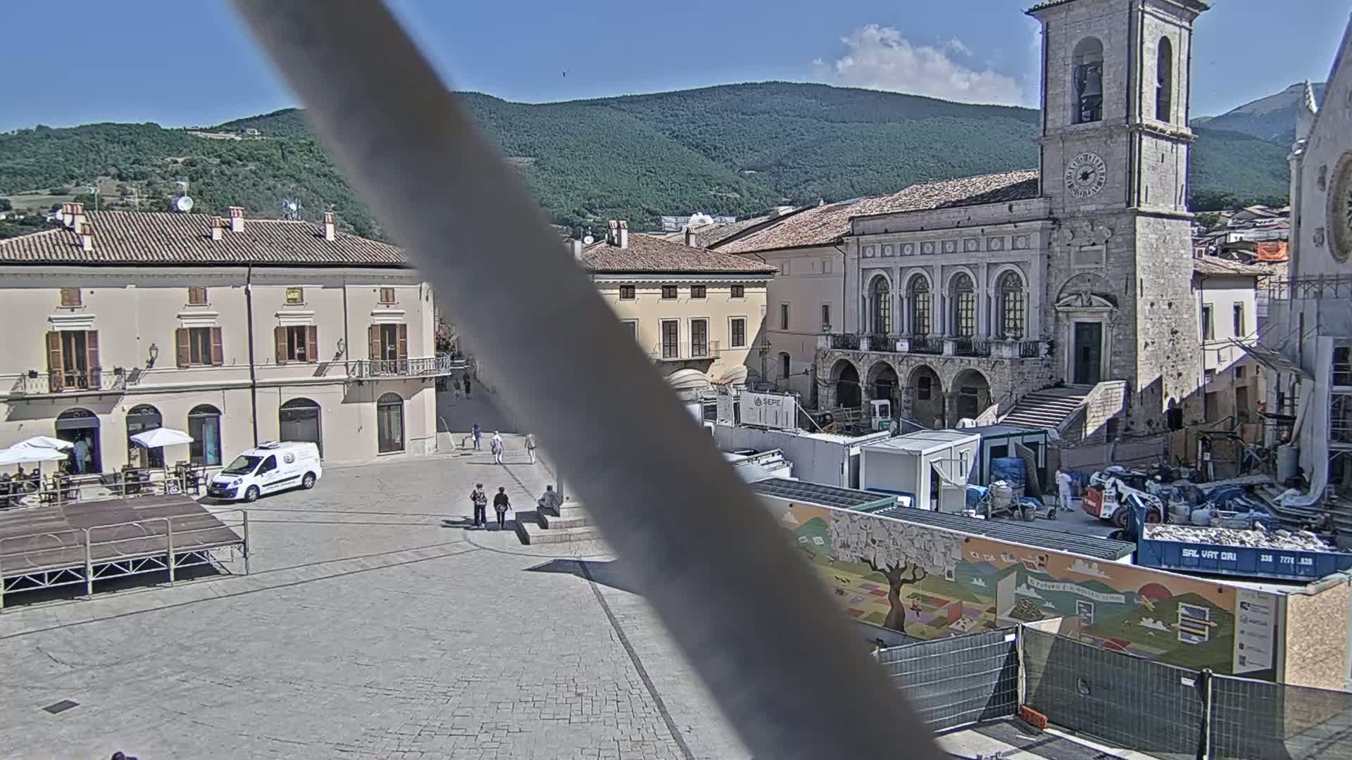 A town square with several buildings, a white van, and a few people, under a mostly sunny sky with mountains in the background.