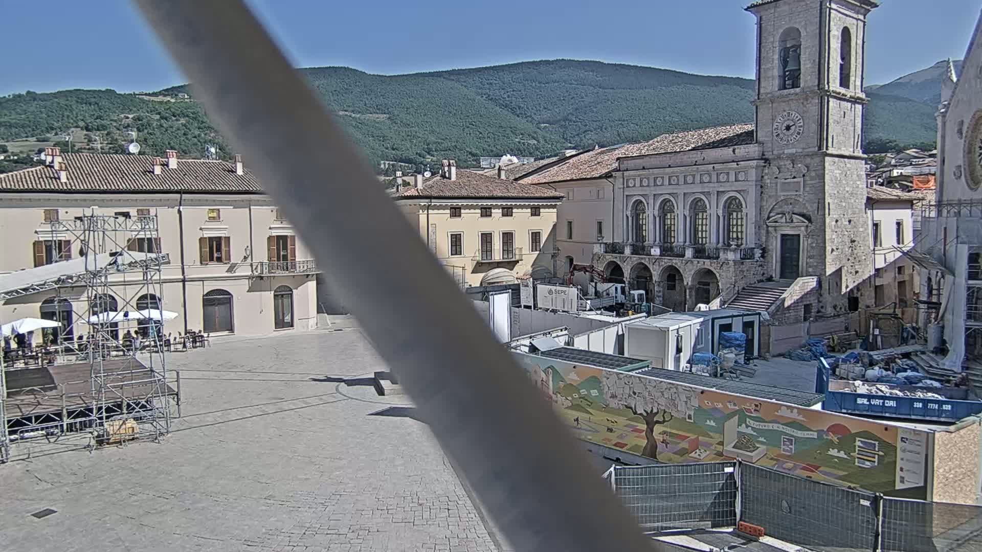 A town square is under construction, with several buildings surrounding it, under a clear sunny sky and mountains in the background.