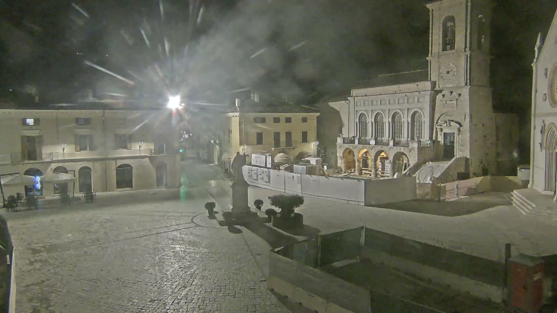 A nighttime view of a historic European town square shows a prominent clock tower, other multi-story buildings, and a section under renovation, all dimly lit under conditions that appear misty or lightly rainy, causing distinct starbursts around the bright streetlights.