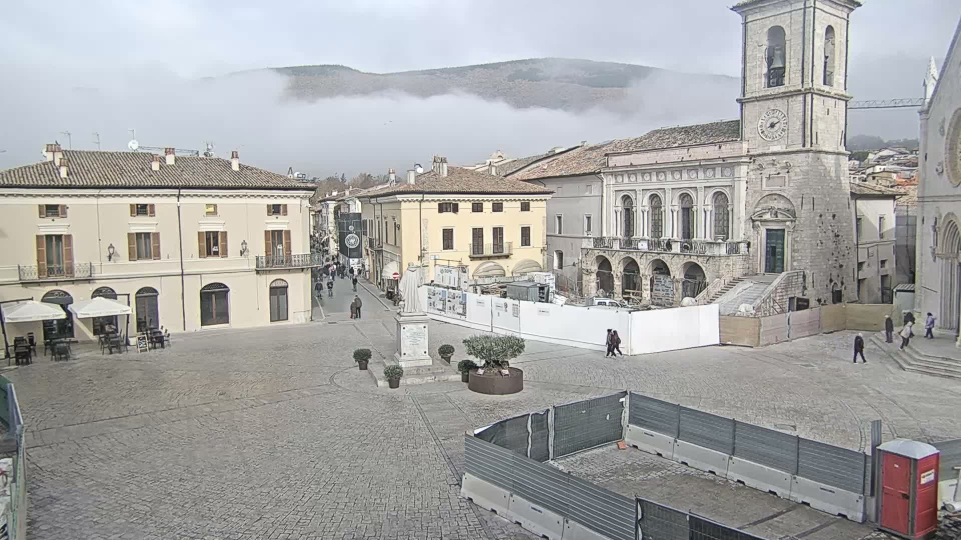 A wide view of a European town square on an overcast day shows historic buildings, including a prominent clock tower, with people walking on the cobblestone ground, some outdoor cafe seating, and distant mountains partially obscured by low clouds.