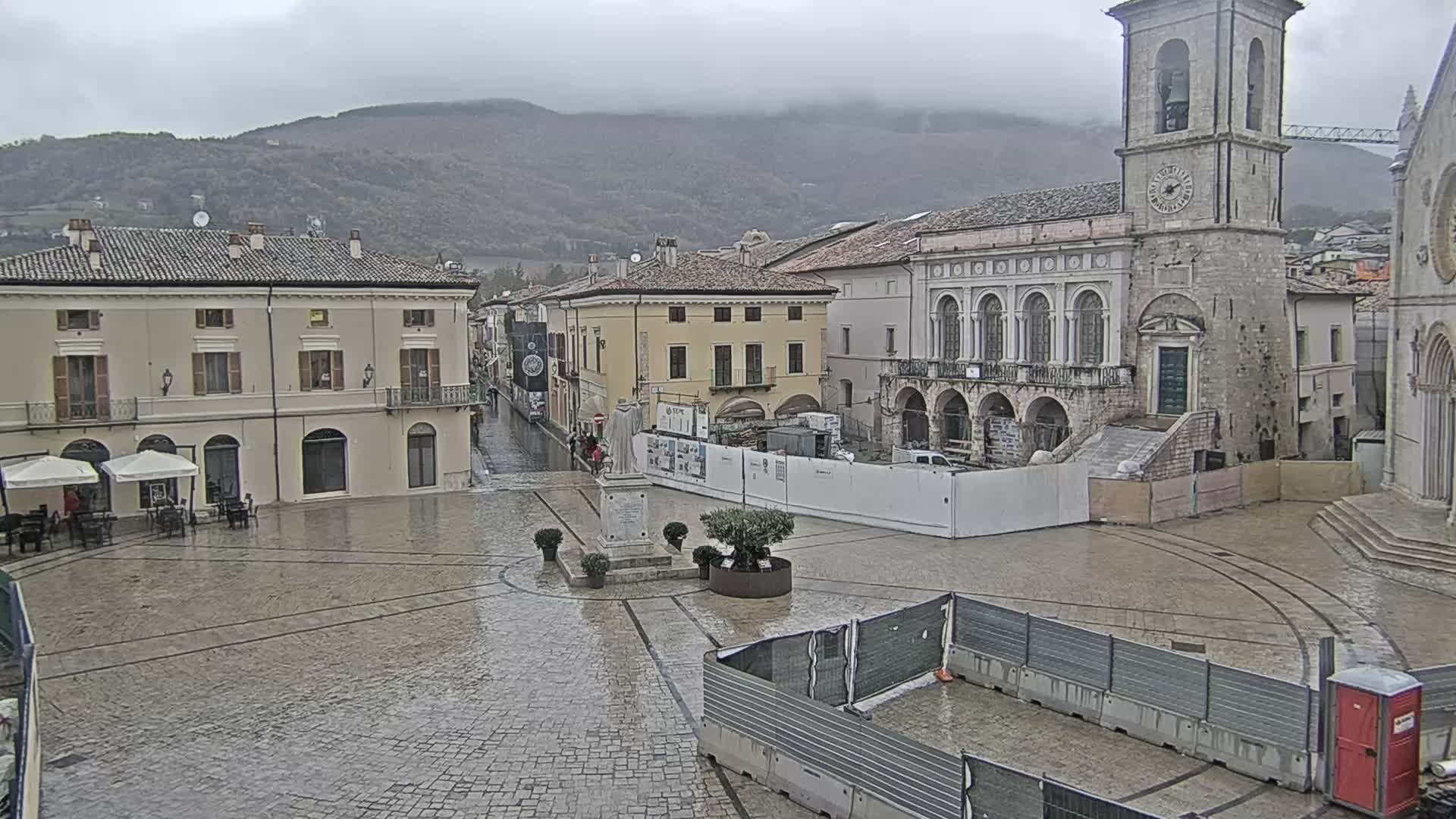 A wide view of a European town square on an overcast day shows historic buildings, including a prominent clock tower, with people walking on the cobblestone ground, some outdoor cafe seating, and distant mountains partially obscured by low clouds.