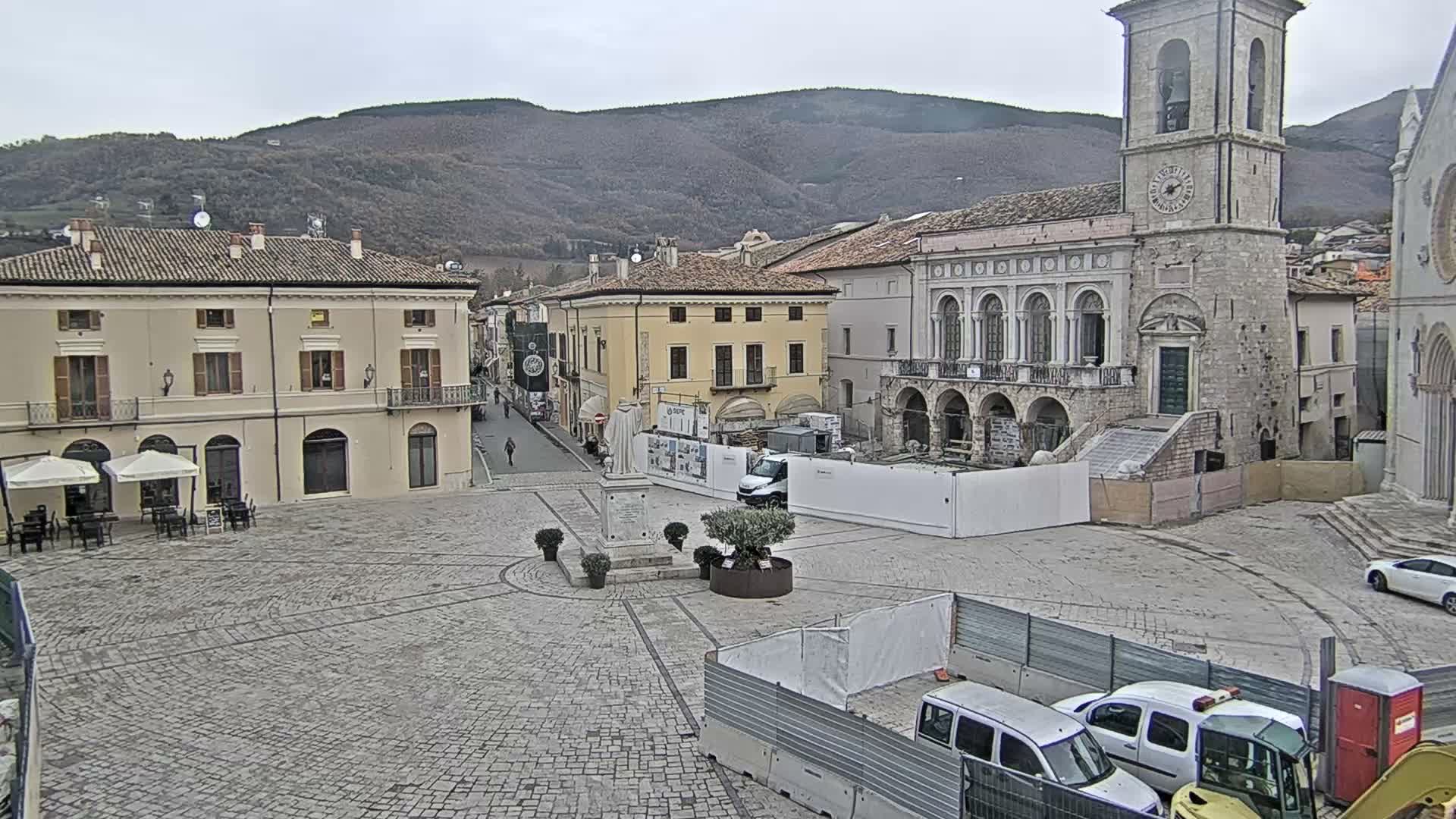A cobblestone town square is visible on an overcast day, framed by historic buildings including a clock tower and a café, with ongoing construction in the foreground and tree-covered mountains in the distance.