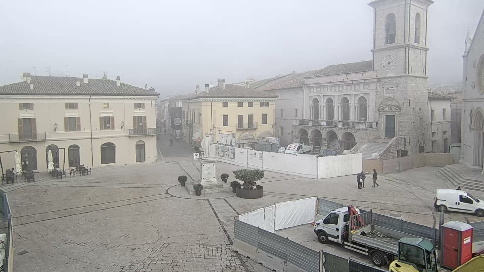 A historic European town square, featuring a prominent clock tower and various buildings, is enveloped in thick fog, with ongoing construction and a few pedestrians and vehicles visible.