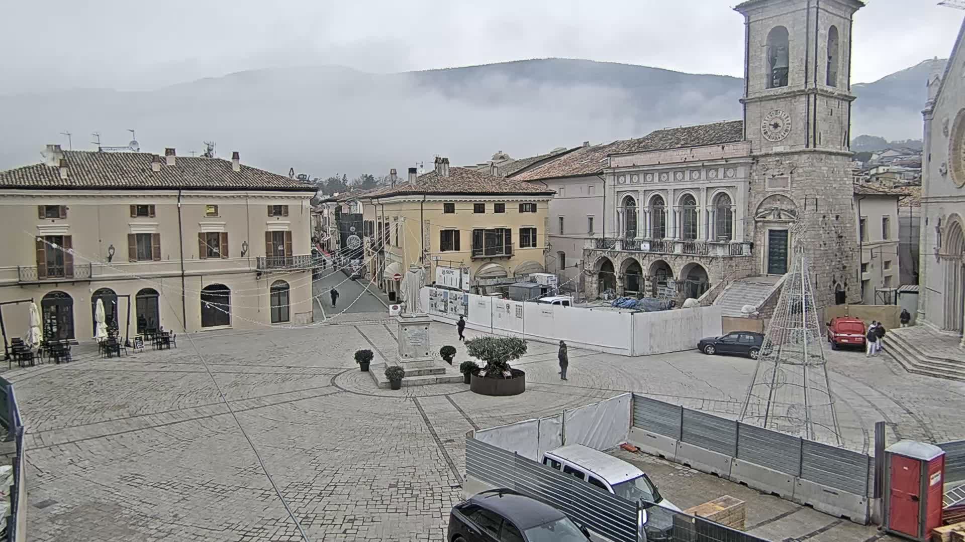 An overcast and misty day casts a subdued light over a historic European town square, which features a prominent stone clock tower, traditional buildings, a central statue, outdoor cafe seating, and scattered pedestrians amidst some construction work, with distant mountains obscured by clouds.