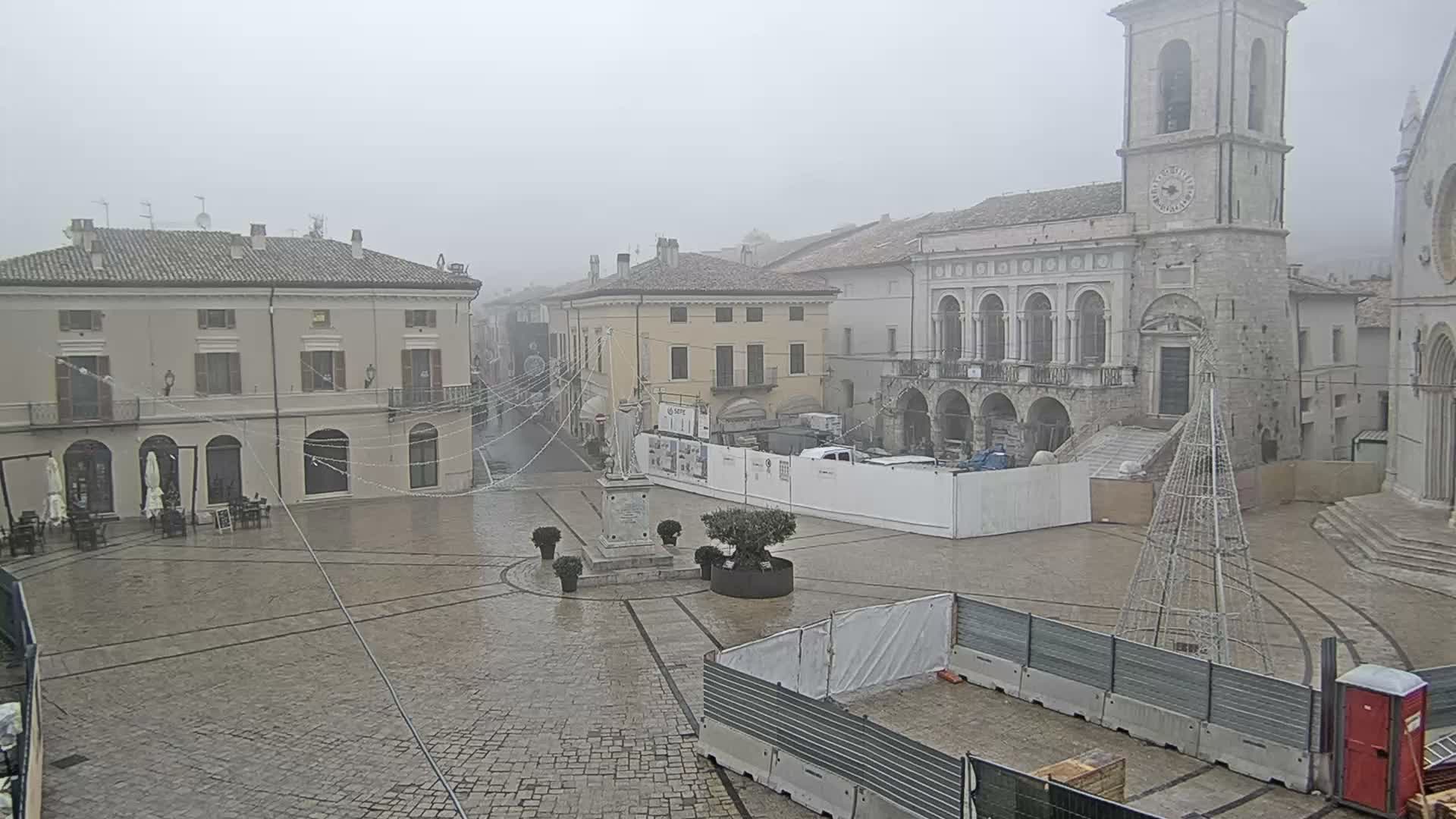 An overcast and misty day casts a subdued light over a historic European town square, which features a prominent stone clock tower, traditional buildings, a central statue, outdoor cafe seating, and scattered pedestrians amidst some construction work, with distant mountains obscured by clouds.