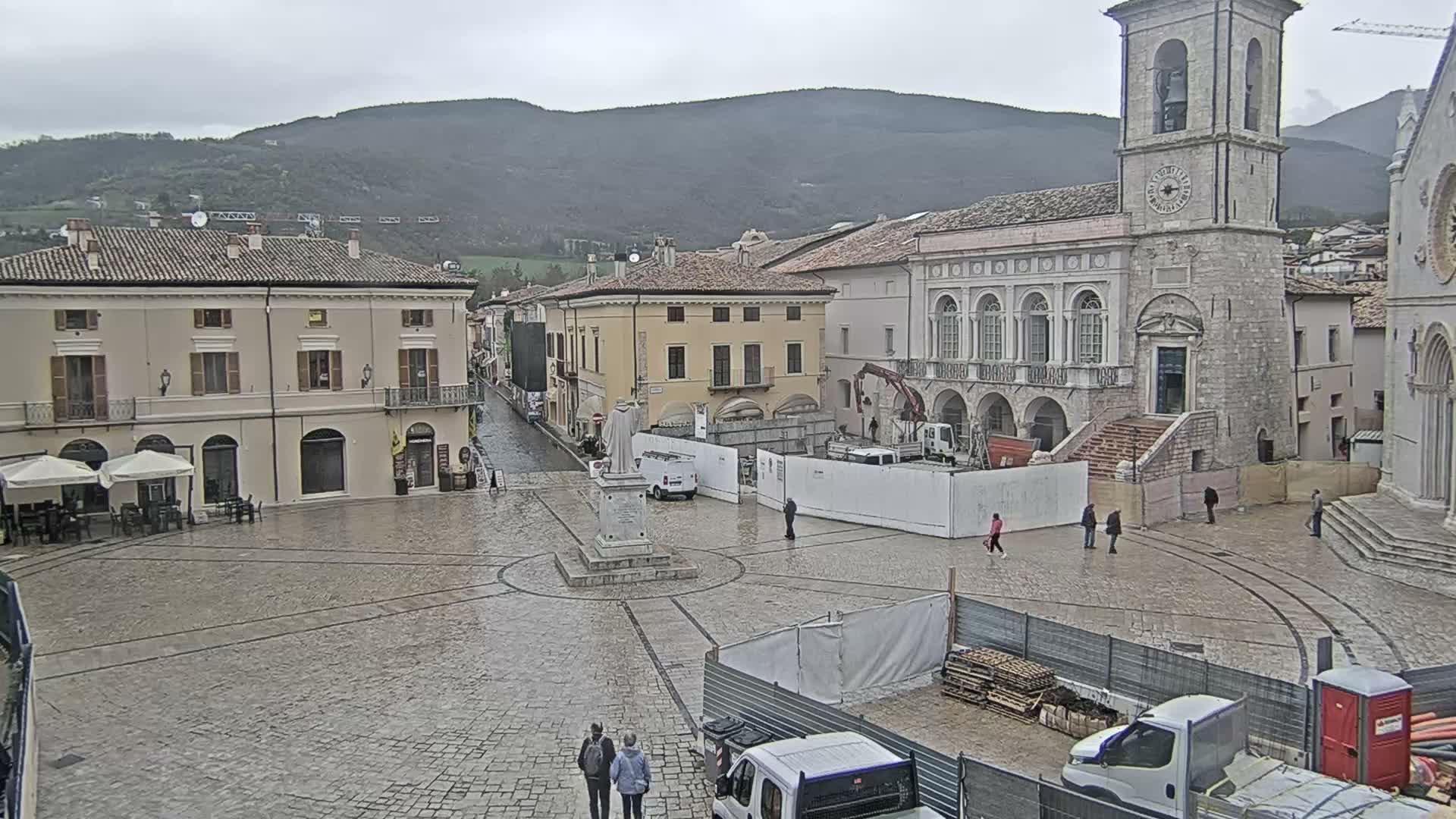 An overcast and misty day casts a subdued light over a historic European town square, which features a prominent stone clock tower, traditional buildings, a central statue, outdoor cafe seating, and scattered pedestrians amidst some construction work, with distant mountains obscured by clouds.