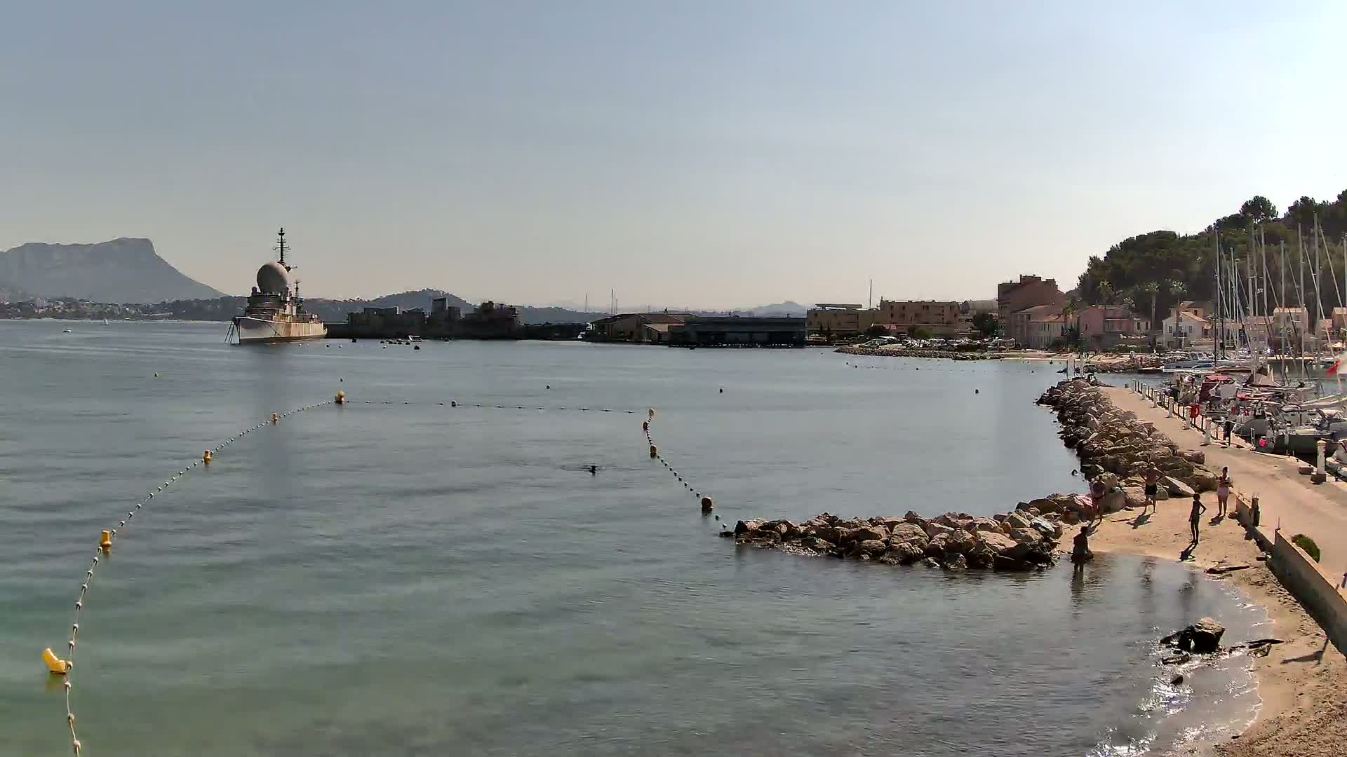 A calm, sunny day shows a harbor with a large ship in the distance, a rocky breakwater leading to a small sandy beach with a few people, and numerous sailboats moored nearby.
