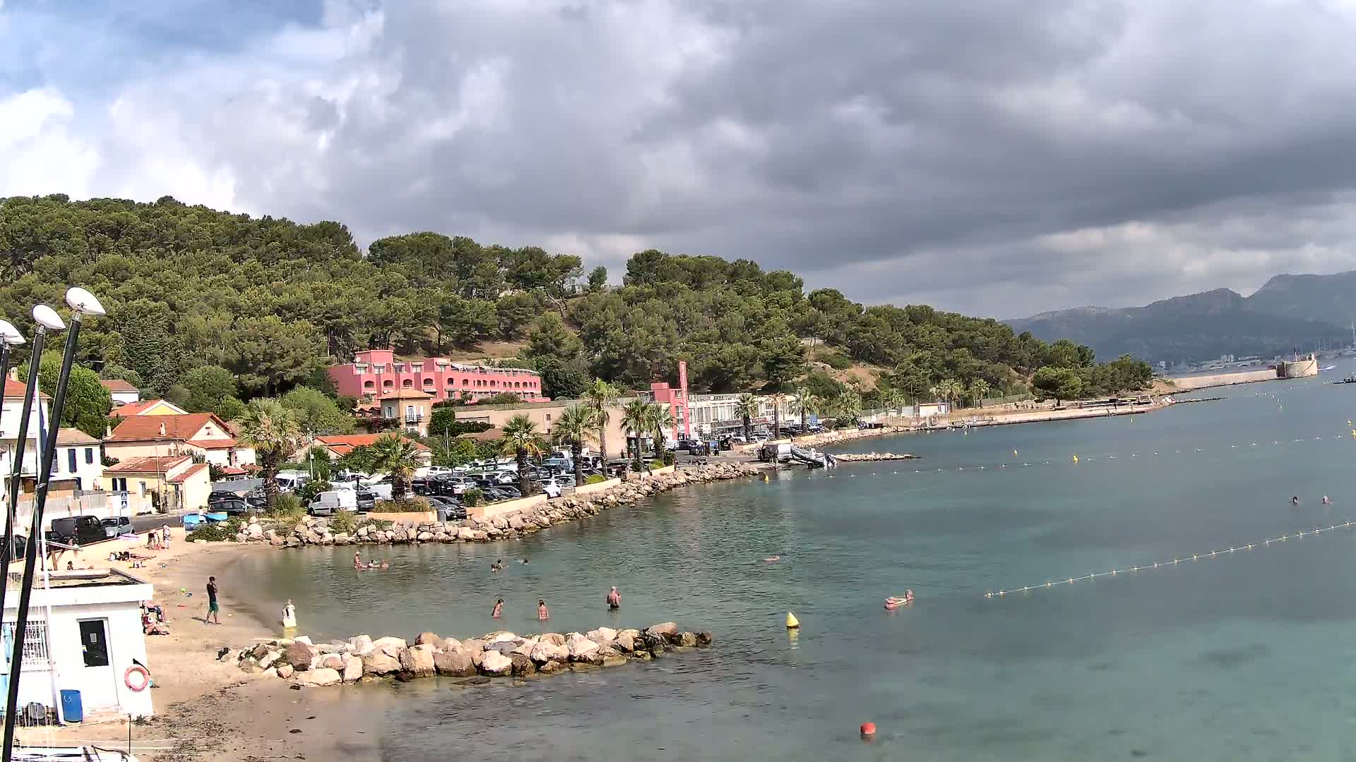 A partly cloudy day at a beach with calm, clear water, people swimming, and buildings along a rocky shore.