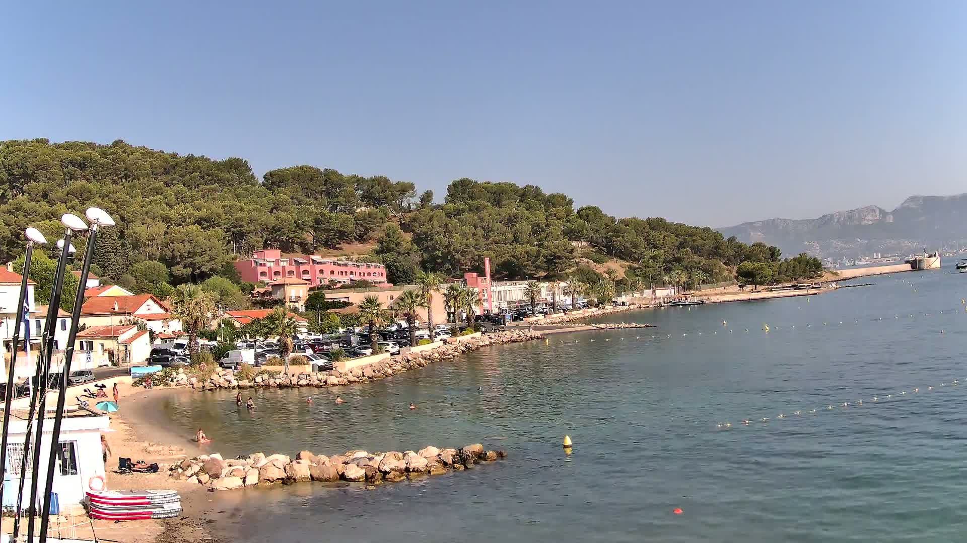 A sunny coastal scene shows a small beach with people swimming, buildings along a shoreline, and a hill covered in green trees, all under a clear blue sky.