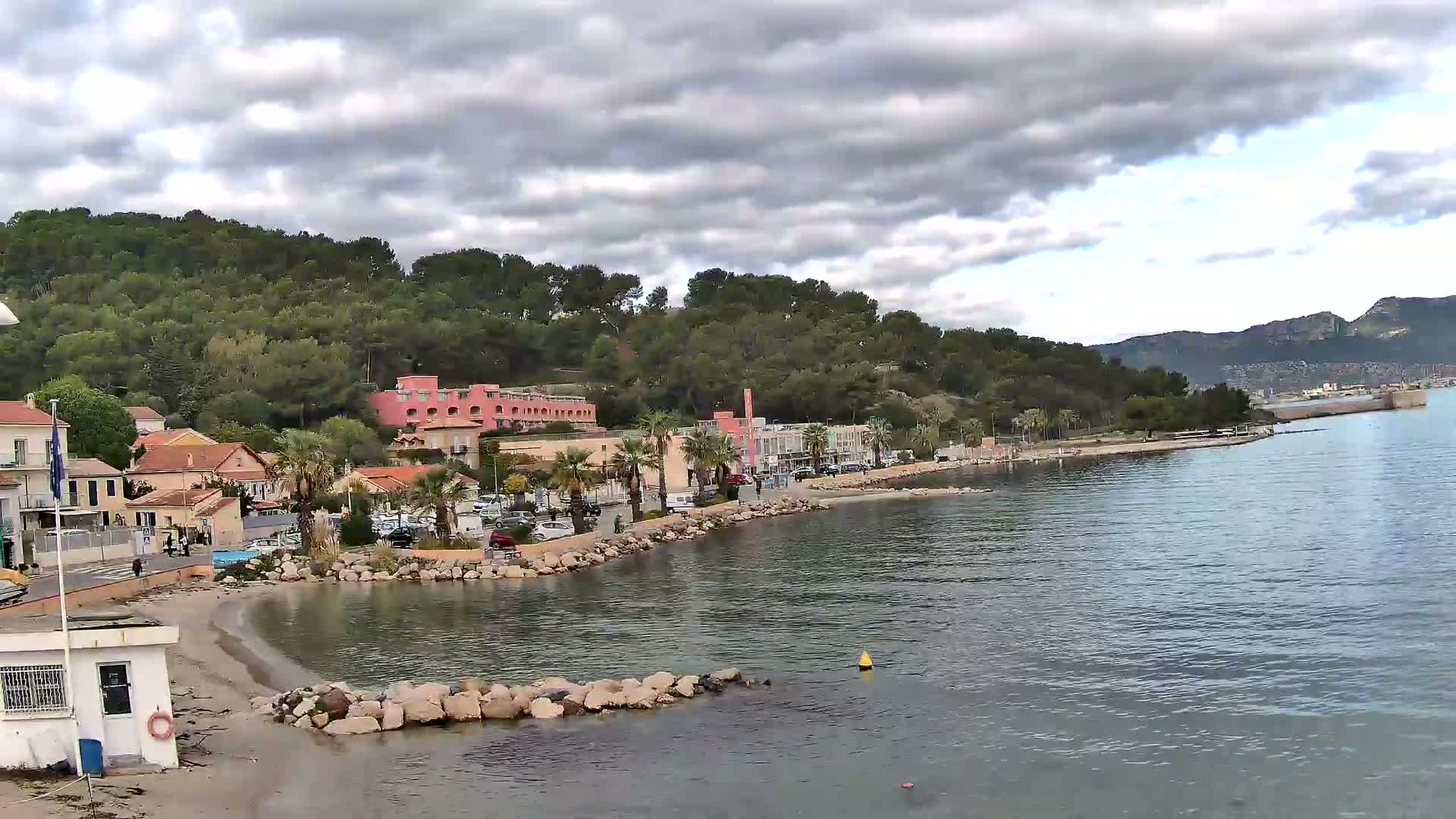 On an overcast day, a coastal town featuring a sandy beach, colorful buildings, and palm trees is nestled at the base of a densely forested hill, overlooking a calm bay that stretches towards distant mountains and a visible industrial port.