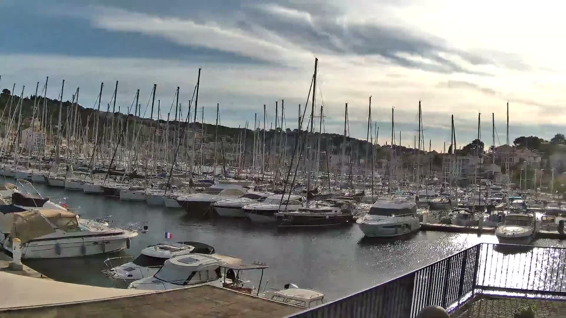 A bustling marina is densely packed with numerous sailboats and motorboats, backed by a hillside town, under a partly cloudy sky with bright sunlight reflecting on the calm water.