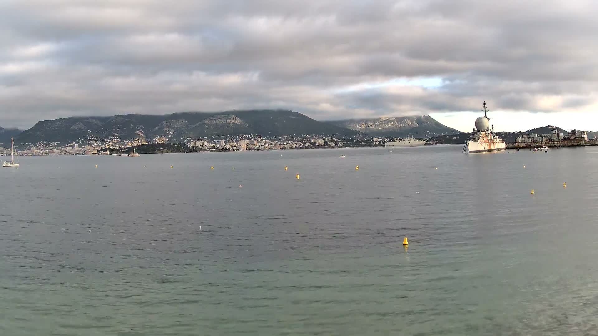Under an overcast sky, a coastal city sprawls along the base of cloud-covered mountains, overlooking a calm bay dotted with yellow buoys, a distant sailboat, and a large ship featuring a prominent spherical radar dome.