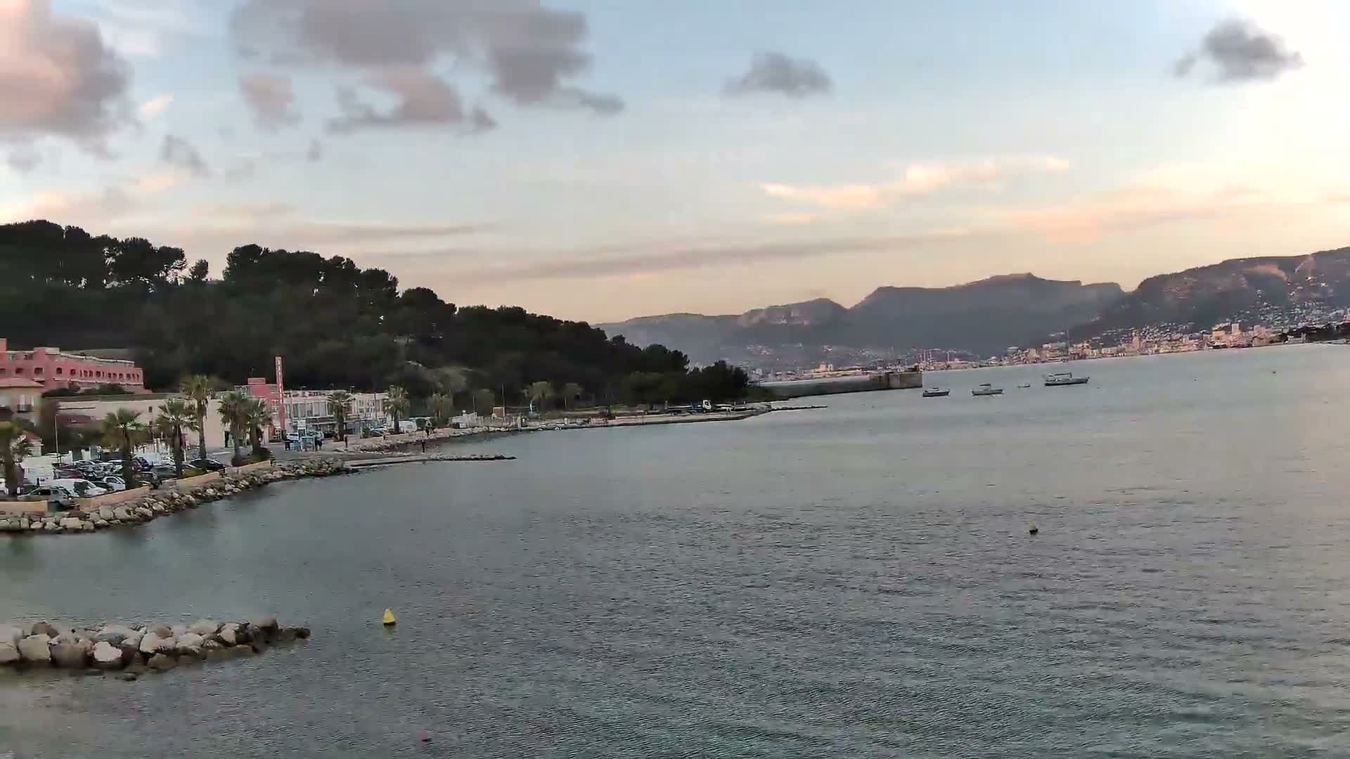 Under an overcast sky, a coastal city sprawls along the base of cloud-covered mountains, overlooking a calm bay dotted with yellow buoys, a distant sailboat, and a large ship featuring a prominent spherical radar dome.